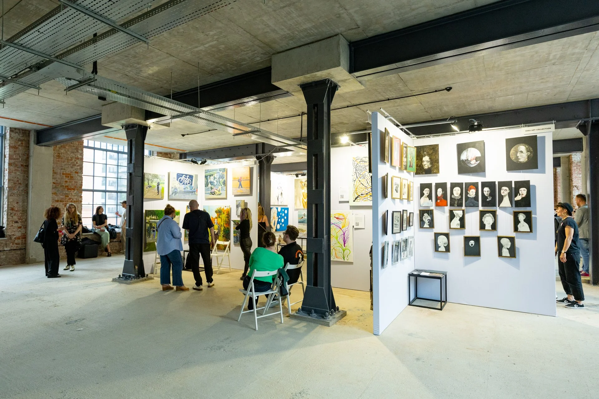 People viewing artwork in an art gallery with exposed brick wall, concrete ceiling, and industrial-style supports.