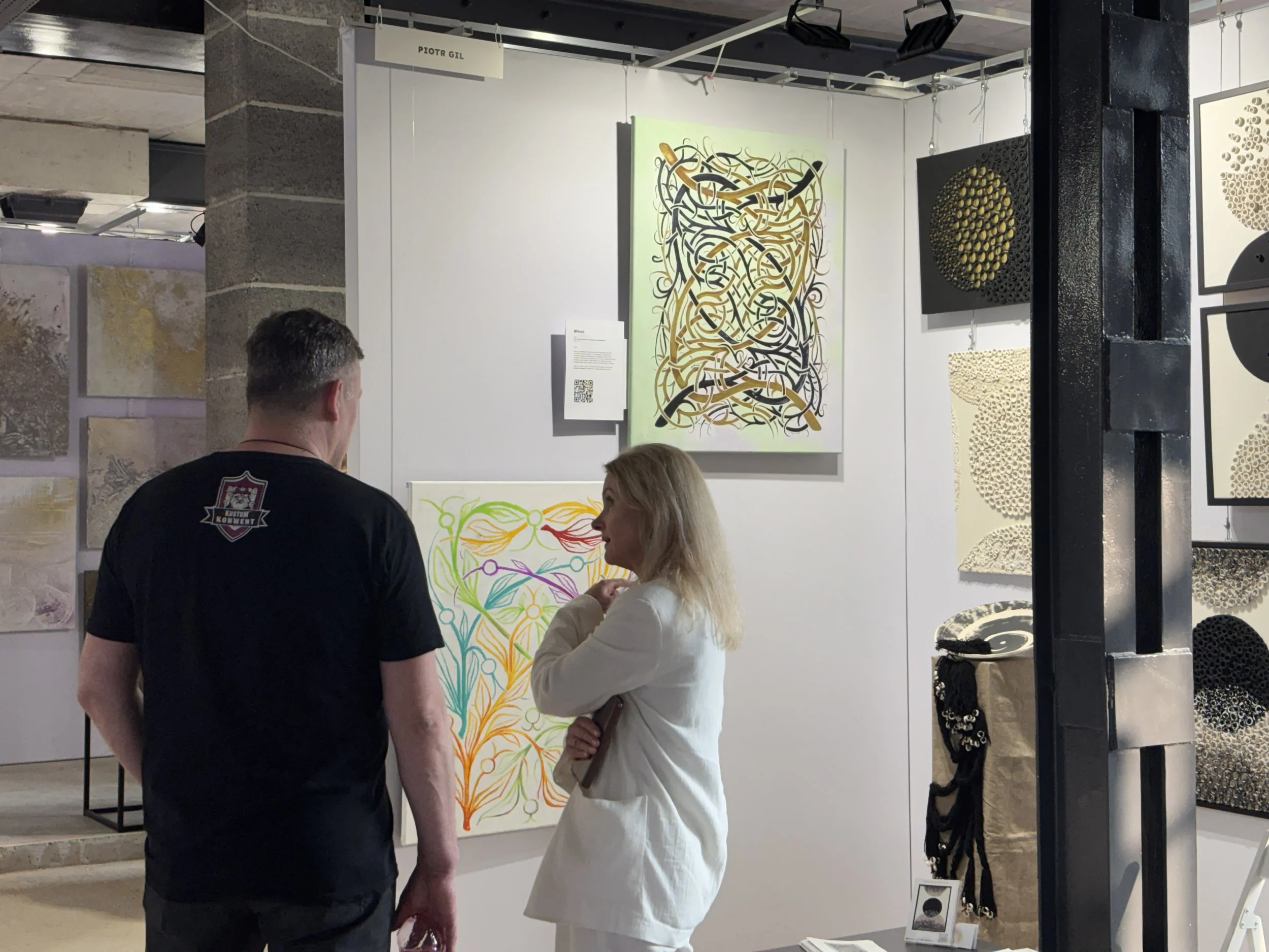 Two people, a man and a woman, stand and converse in an art gallery. The man faces away and has dark hair, while the woman has blonde hair. Behind them is an abstract painting with swirls in black, gold, and white, and other artwork and display items are visible on the gallery walls.