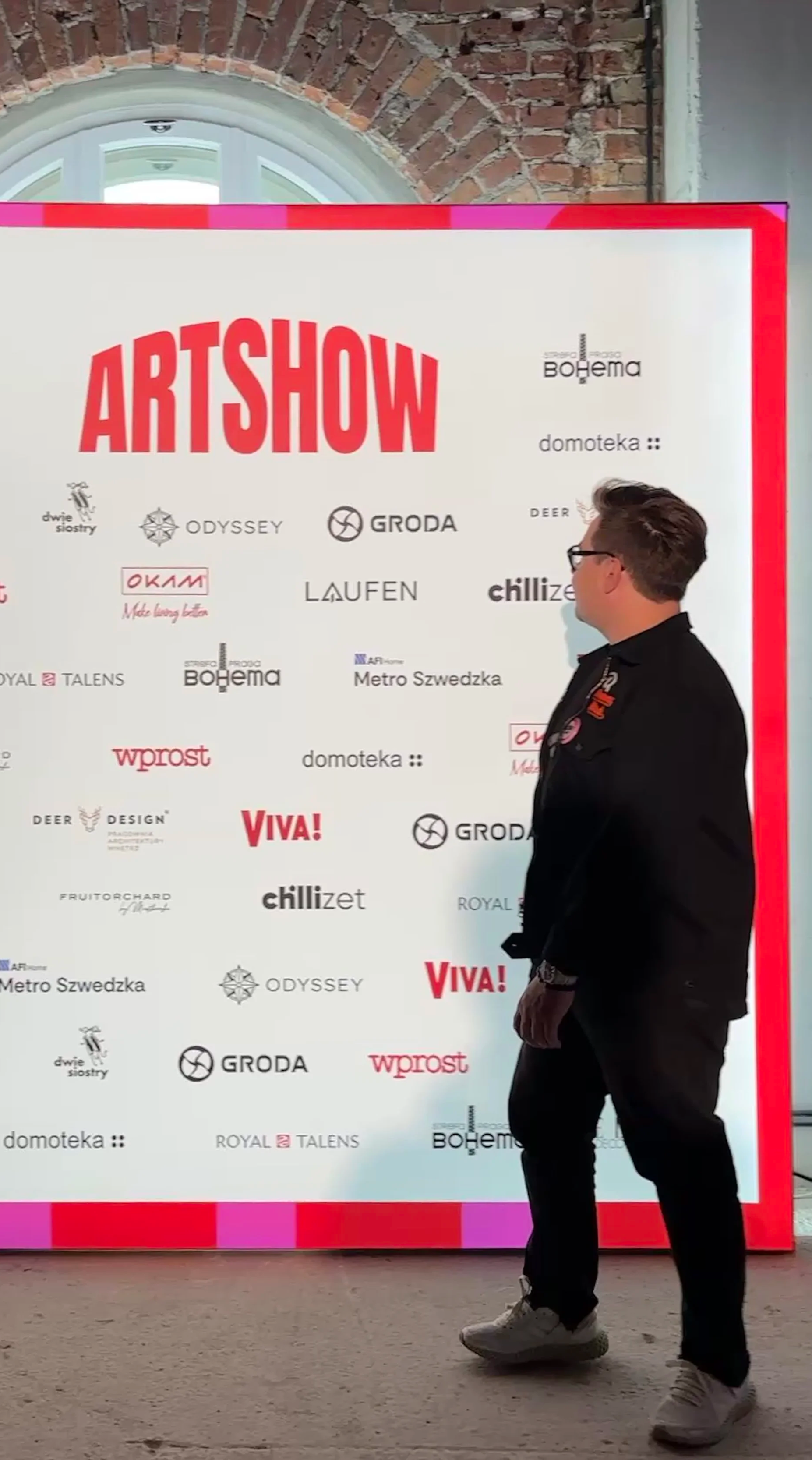 Man standing in front of a large white poster with red and black text, attending an art show. The poster displays the word 'ARTSHOW' in large red letters and features various logos.