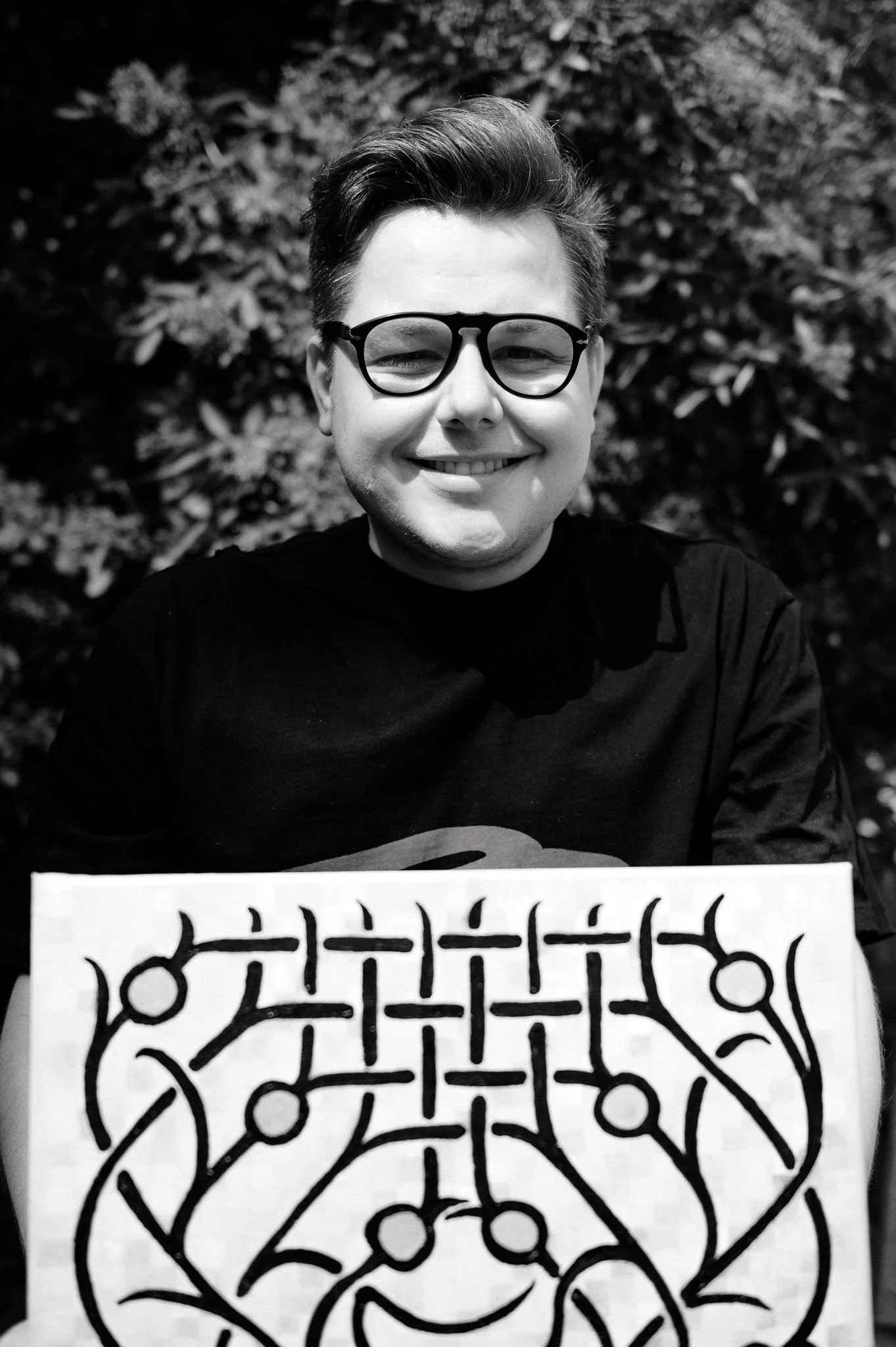 A smiling person Peter Gil with short hair and glasses, holding the "Generations" painting with abstract black lines, posing outdoors against a leafy background.