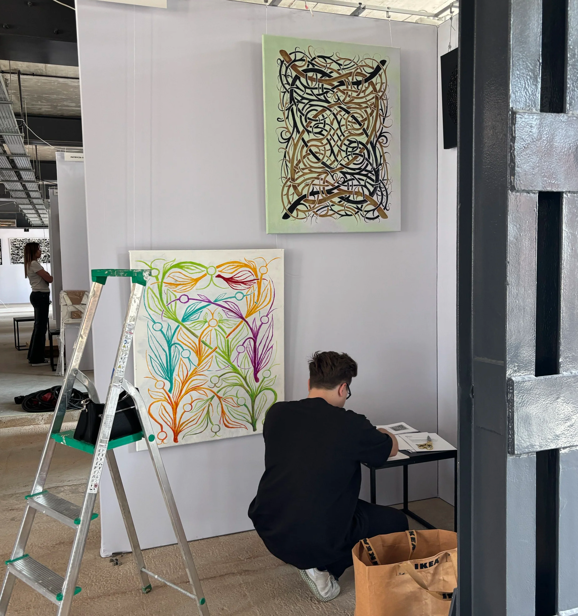 A man with dark hair and glasses sitting at a table inside an art display area, wearing a black shirt. Behind him is a colorful artwork with abstract, flowing lines in rainbow colors. To the left, a woman with long hair and wearing a white top is standing and looking at the art.