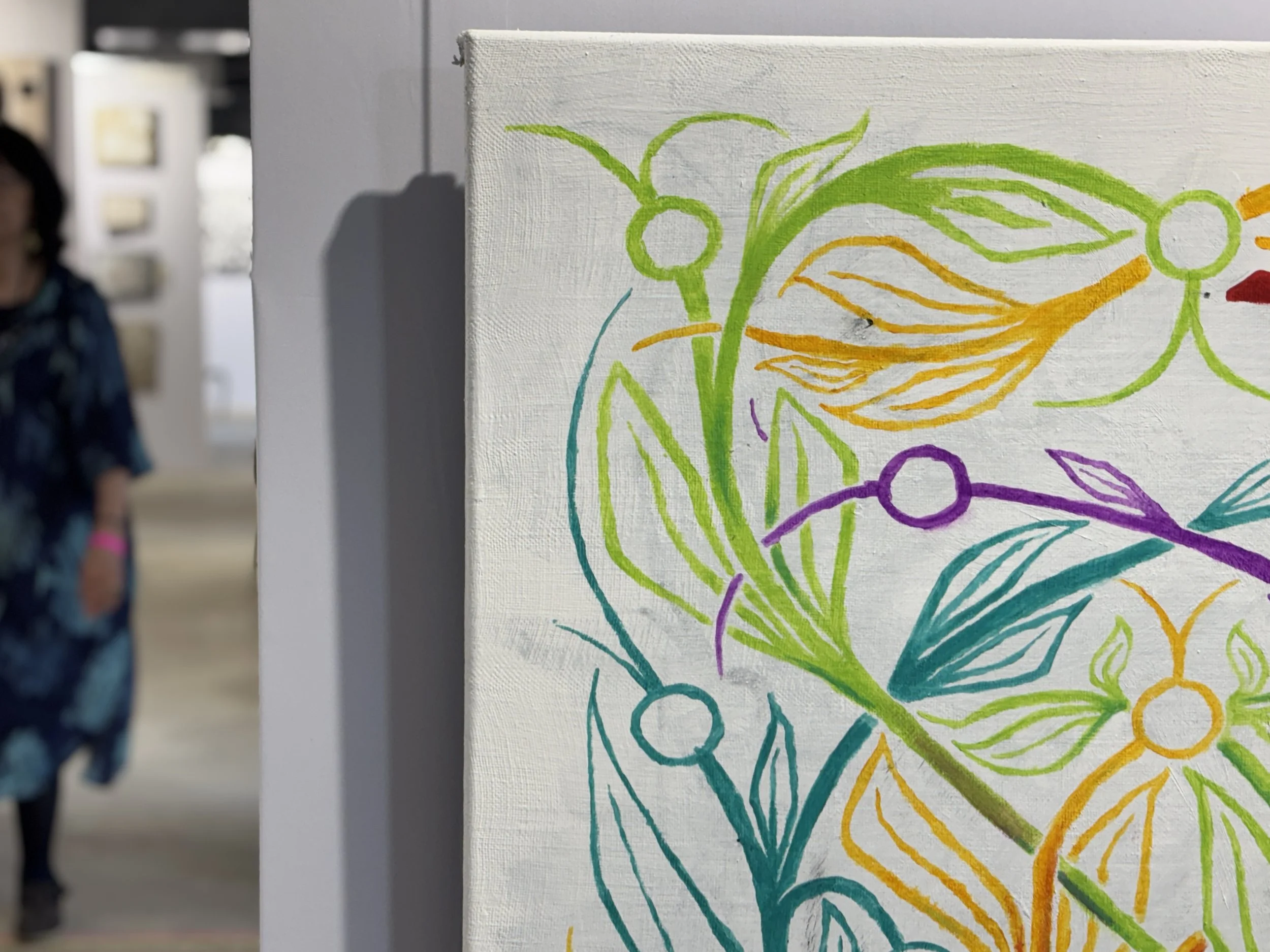 A colorful, abstract painting of swirling lines and shapes in green, yellow, purple, turquoise, and red, displayed on a white canvas in an indoor gallery setting. A blurred person is visible in the background.