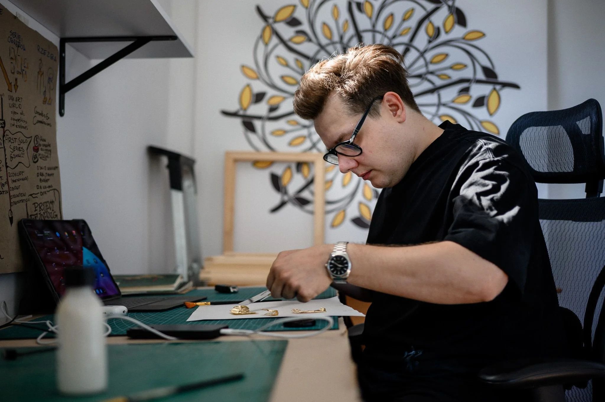 Peter Gil working with gold leaf at a desk in an his art studio. In the background, a decorative wall art.