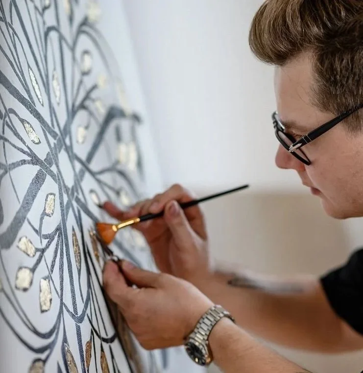 A person with glasses painting a detailed, artistic wall design with a small brush.