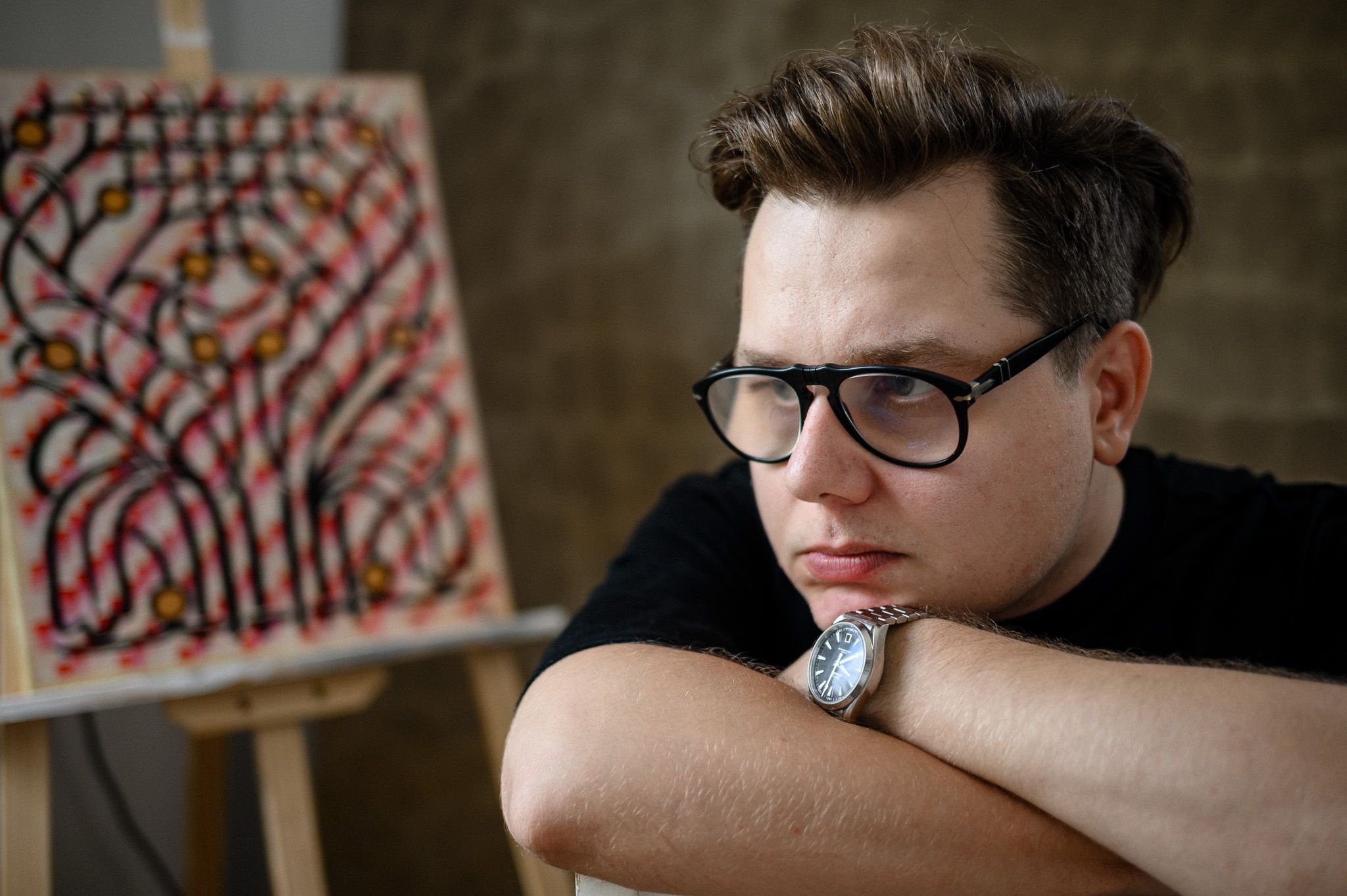 A young man with short hair (Peter Gil) wearing black-rimmed glasses and a silver watch on his left wrist, resting his head on crossed arms. In the background, there is a blurred abstract painting with black, red, and yellow colors.