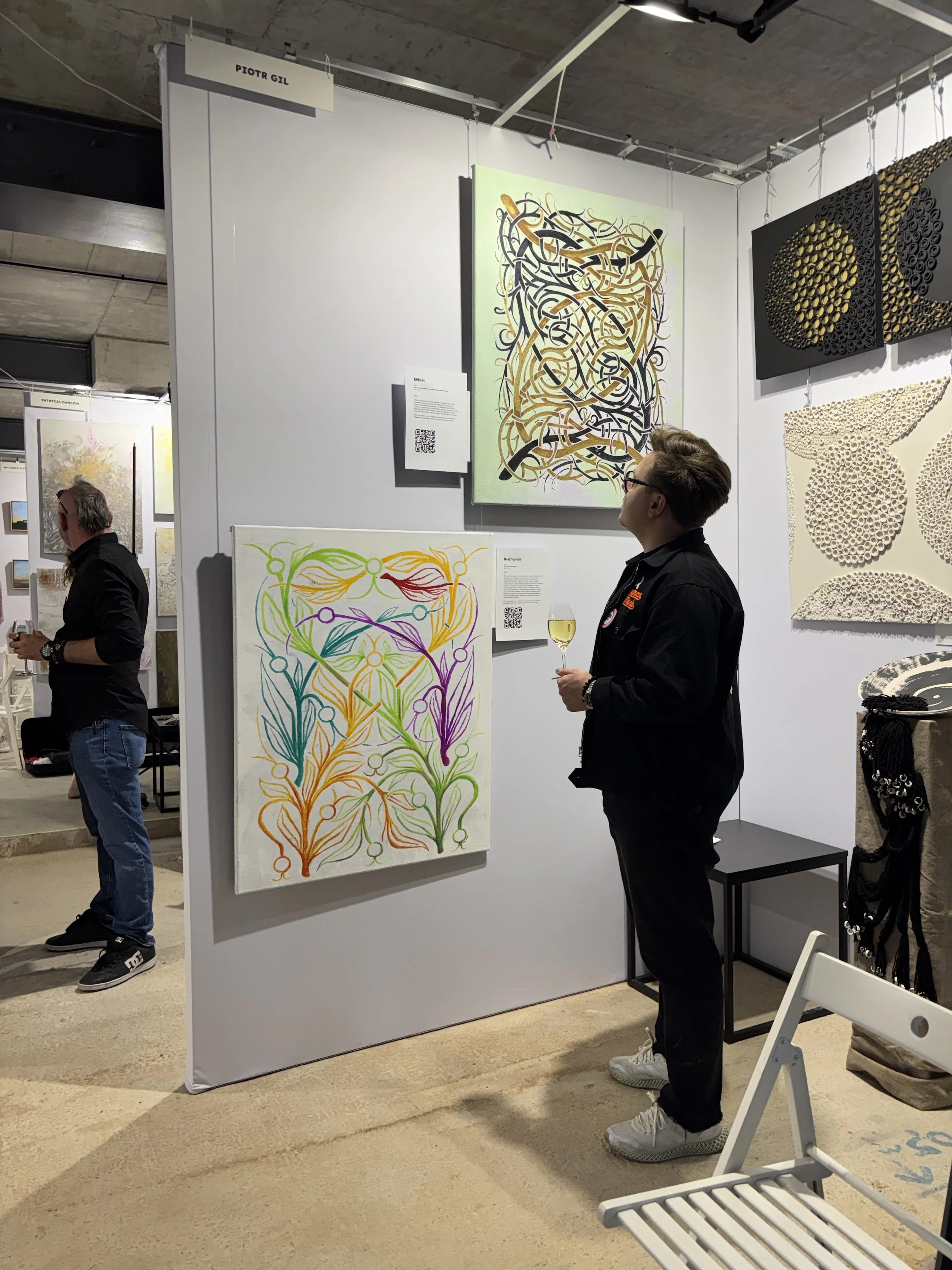 An art exhibition with abstract paintings on a wall, including a colorful piece with intertwined lines and a black-and-golden panel with circular patterns. Two men are viewing the artwork, one holding a glass of white wine.