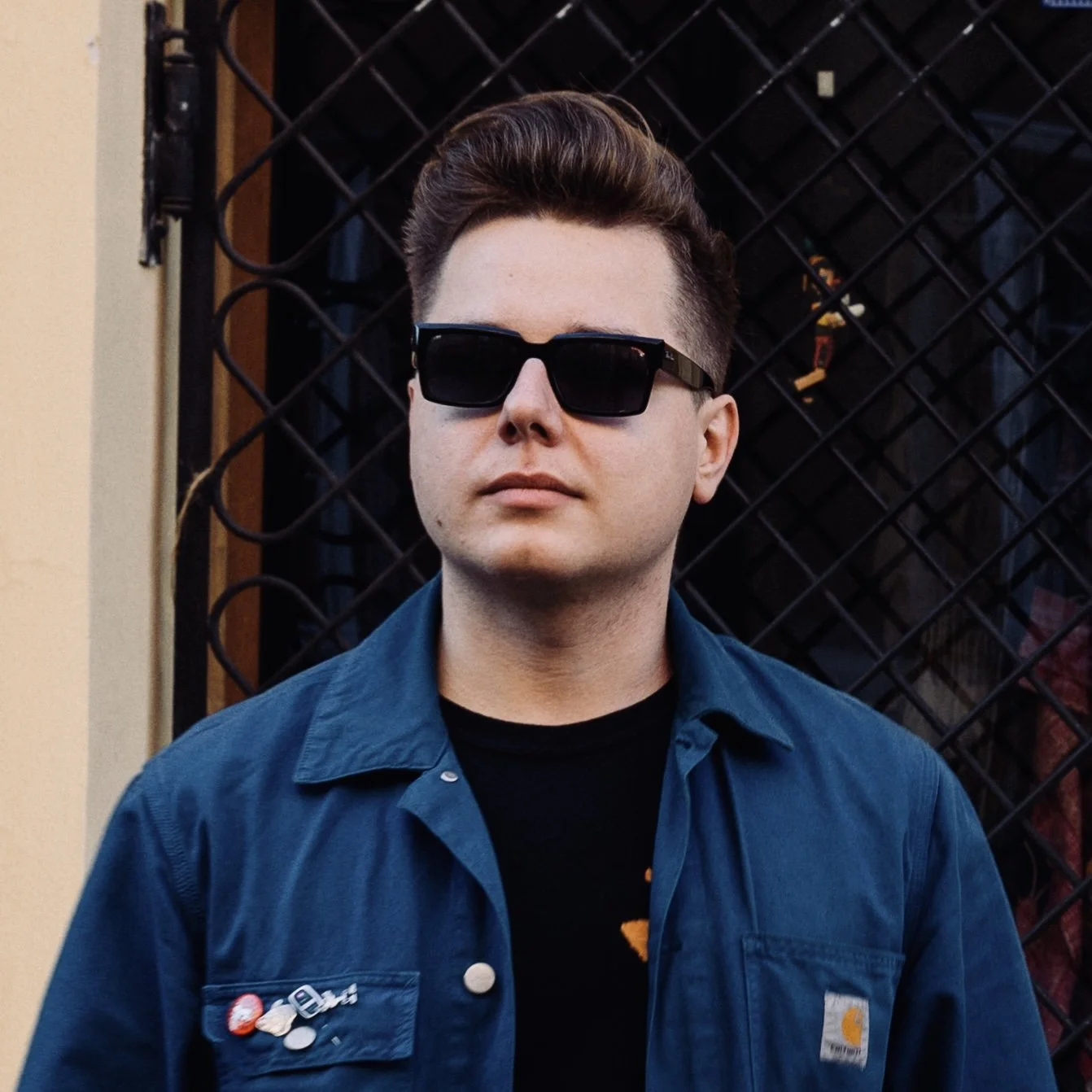 Young man with styled hair wearing black sunglasses, a blue jacket with pins, and a black shirt, standing in front of a black metal fence.