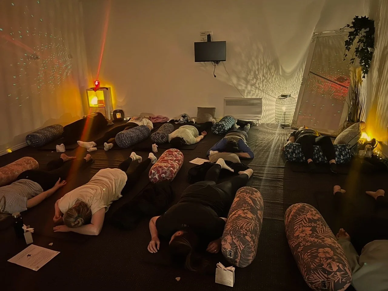 People lying on their stomachs with cushions in a dimly lit room, possibly practicing yoga or meditation.