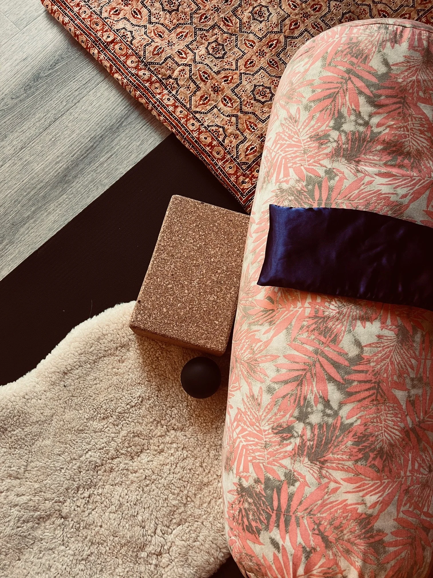 Top view of a corner with a beige and gray patterned sofa, a navy blue cushion, a pink and gray floral patterned pillow, a small granite side table, a black round object, a beige textured rug, a dark brown surface, and a red and blue patterned rug.