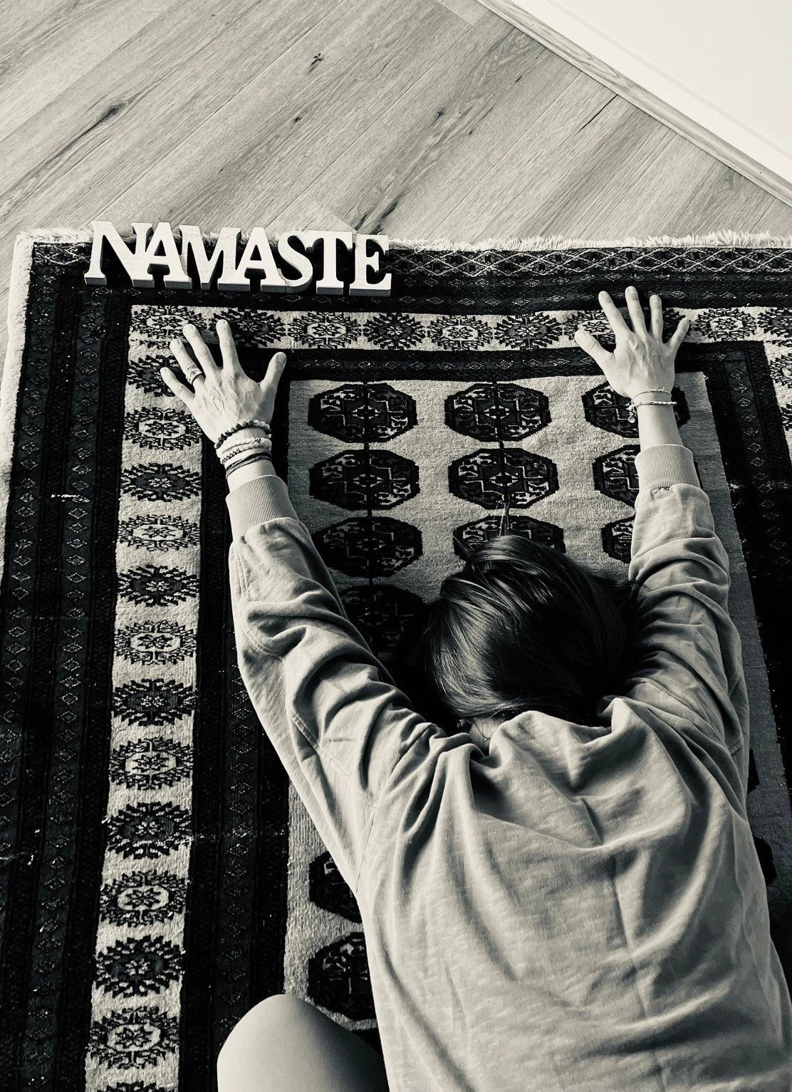 A person is lying on a patterned rug with arms stretched upward. The person is wearing a long-sleeved shirt, bracelets, and has a calm demeanor. The word 'NAMASTE' is placed at the top left corner of the rug.