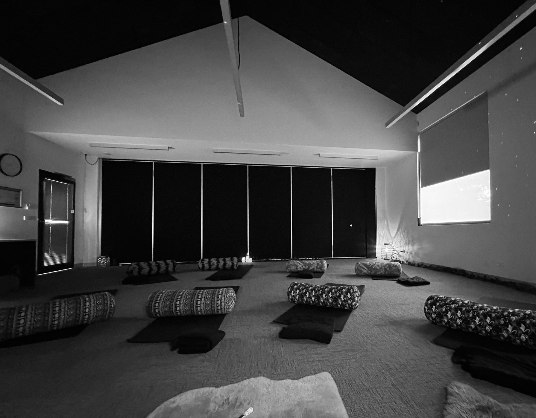 A dark room with yoga mats and bolsters arranged on the floor, large windows with blinds, and black blinds covering the back wall.