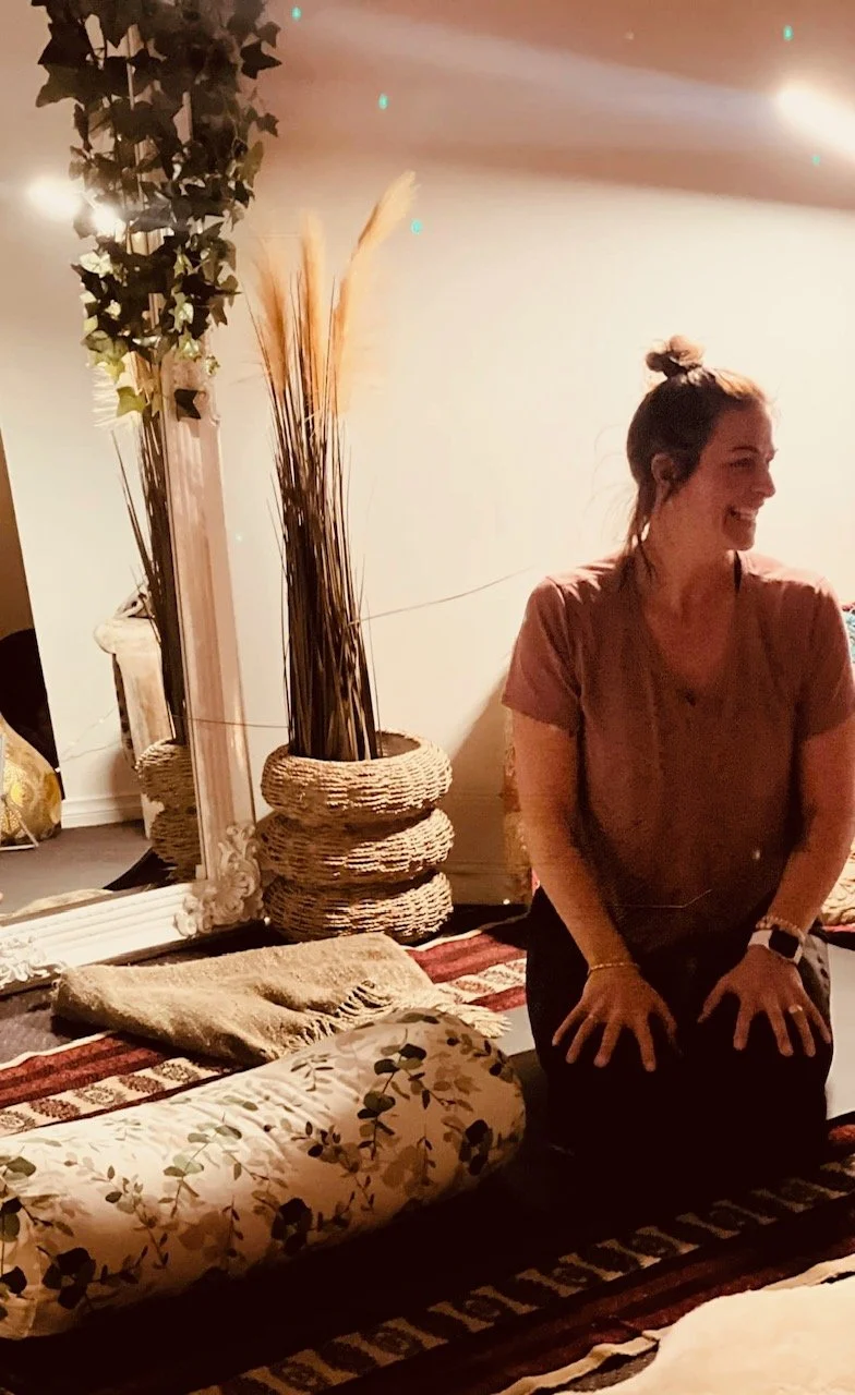 A woman with a bun hairstyle, wearing a brown shirt, sits on her knees on a patterned rug in a room decorated with plants, wicker baskets, a mirror, and cushions, smiling and looking to the right.