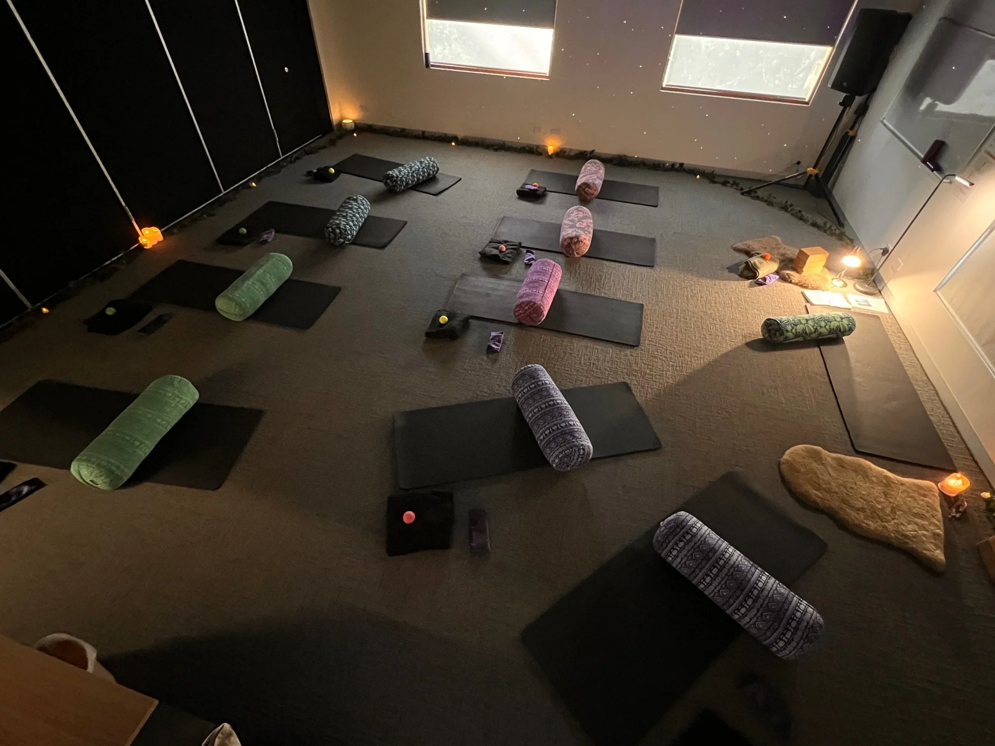 Yoga mats with bolsters and props arranged for a class in a dimly lit room with large windows.