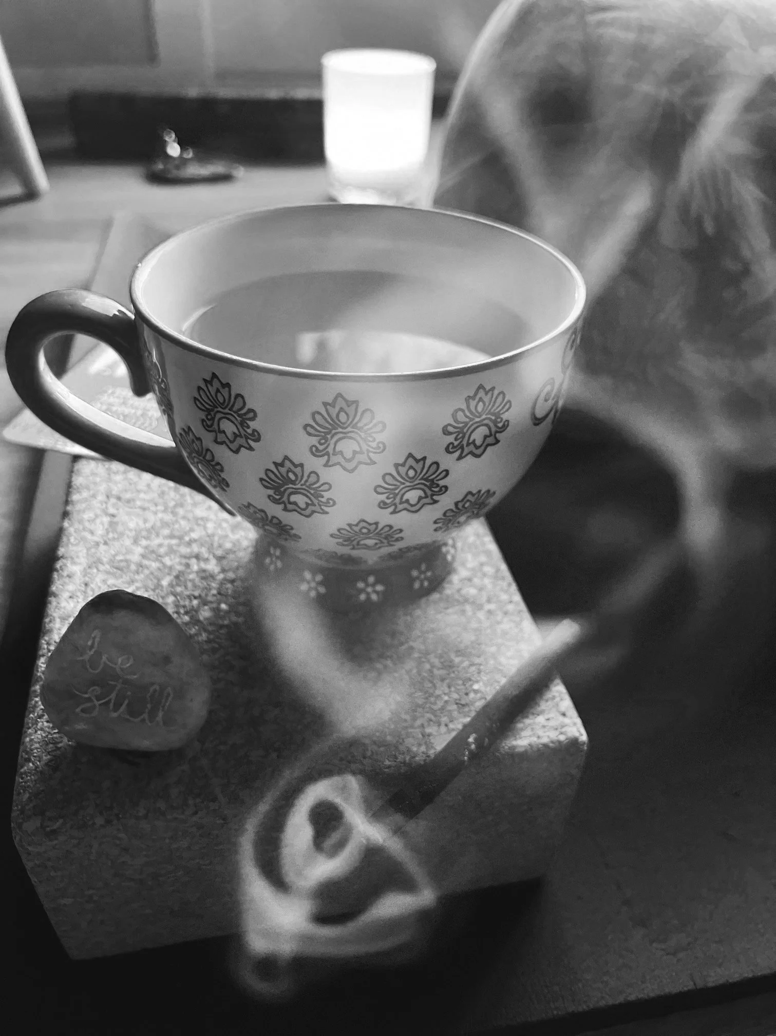 A cup of hot beverage on a kitchen table with a decorative heart-shaped stone that says "be still", and a spoon resting on the surface. In the background, there is a glass filled with water and some household items.