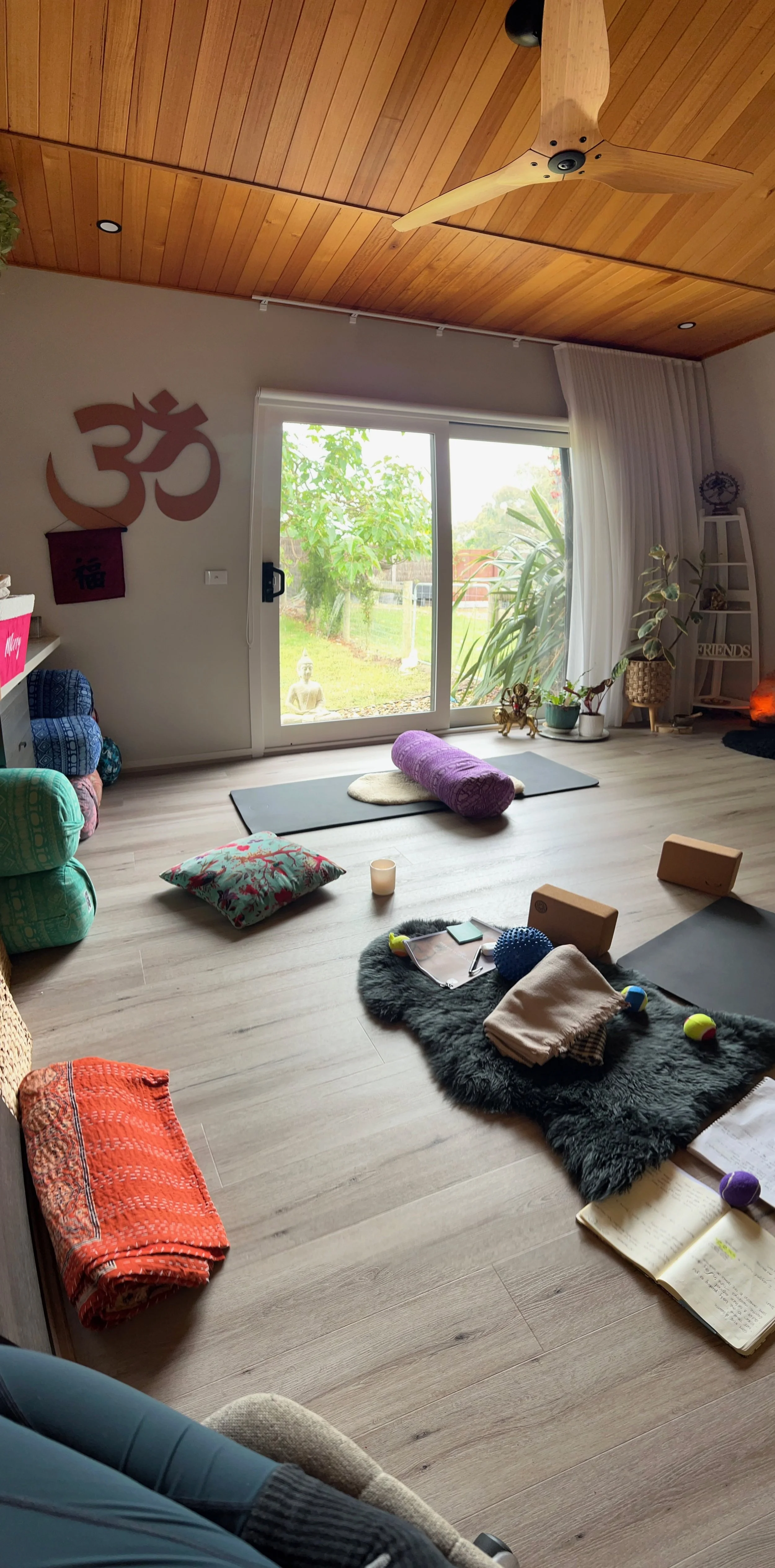 Interior of a yoga or meditation studio with a large glass door leading outside, wooden ceiling with ceiling fan, yoga mats, pillows, candles, and various decorations on the floor and walls.