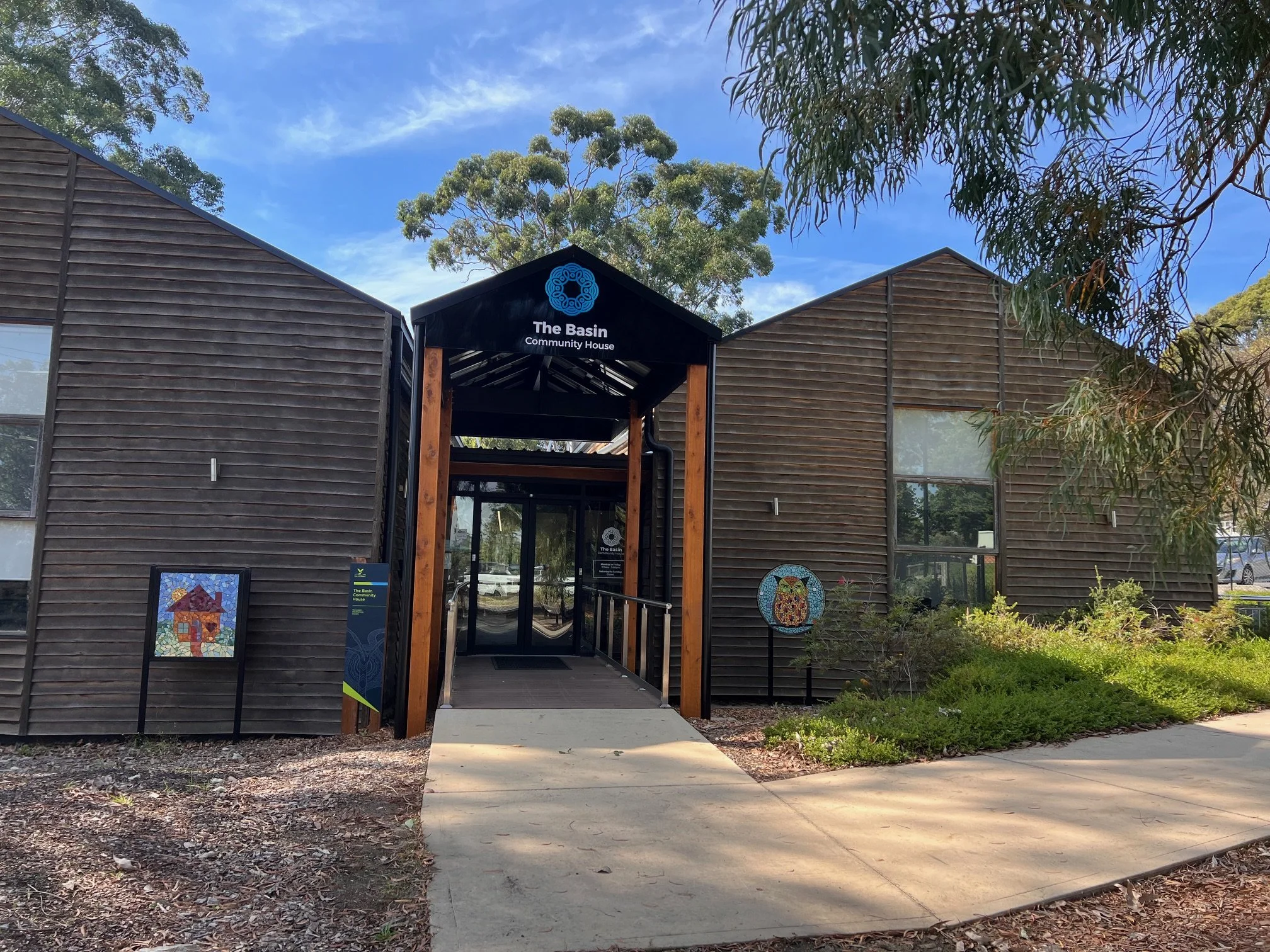 The Basin Community House, a modern building with dark wooden exterior, has an entrance with a black gabled roof and wooden supports, surrounded by greenery and trees under a blue sky.