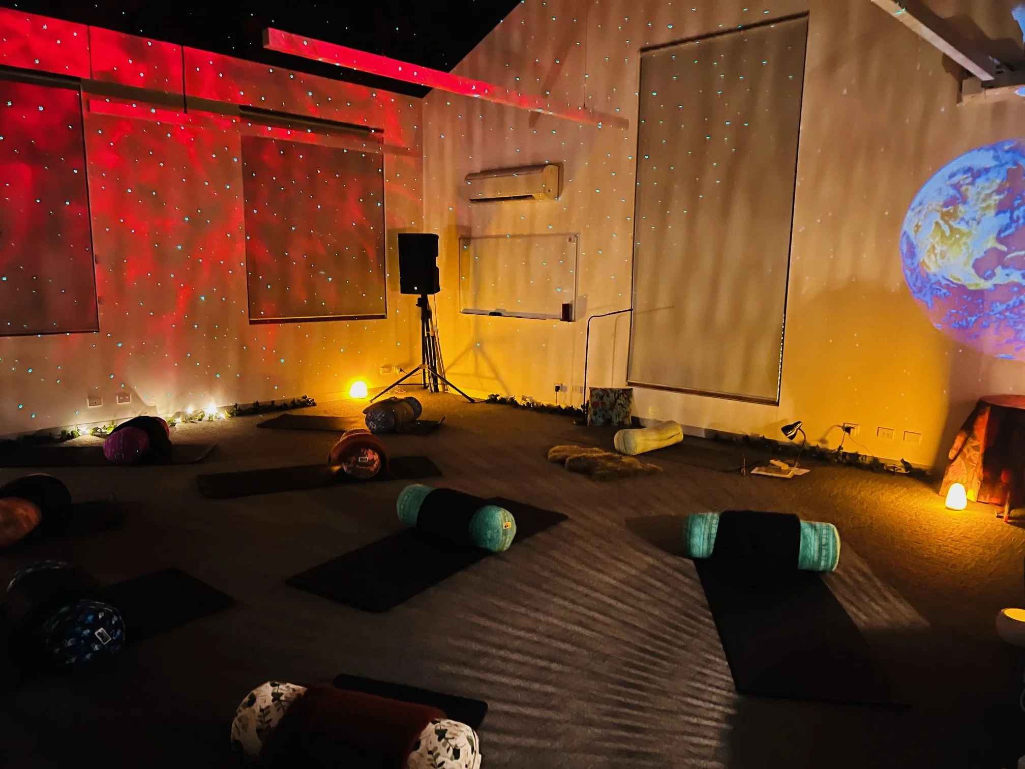Room decorated for a yoga or meditation session with mats, bolsters, and props, a large projection of Earth on the wall, and colorful lights creating a cozy atmosphere.