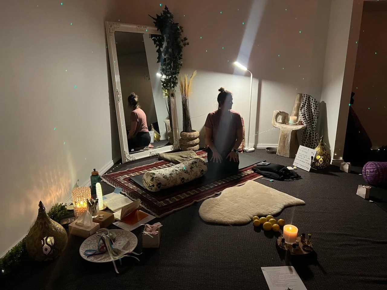 A woman kneels on a rug with her eyes closed in a dimly lit room decorated with candles, cushions, a mirror, and room decor, performing a meditation or spiritual practice.