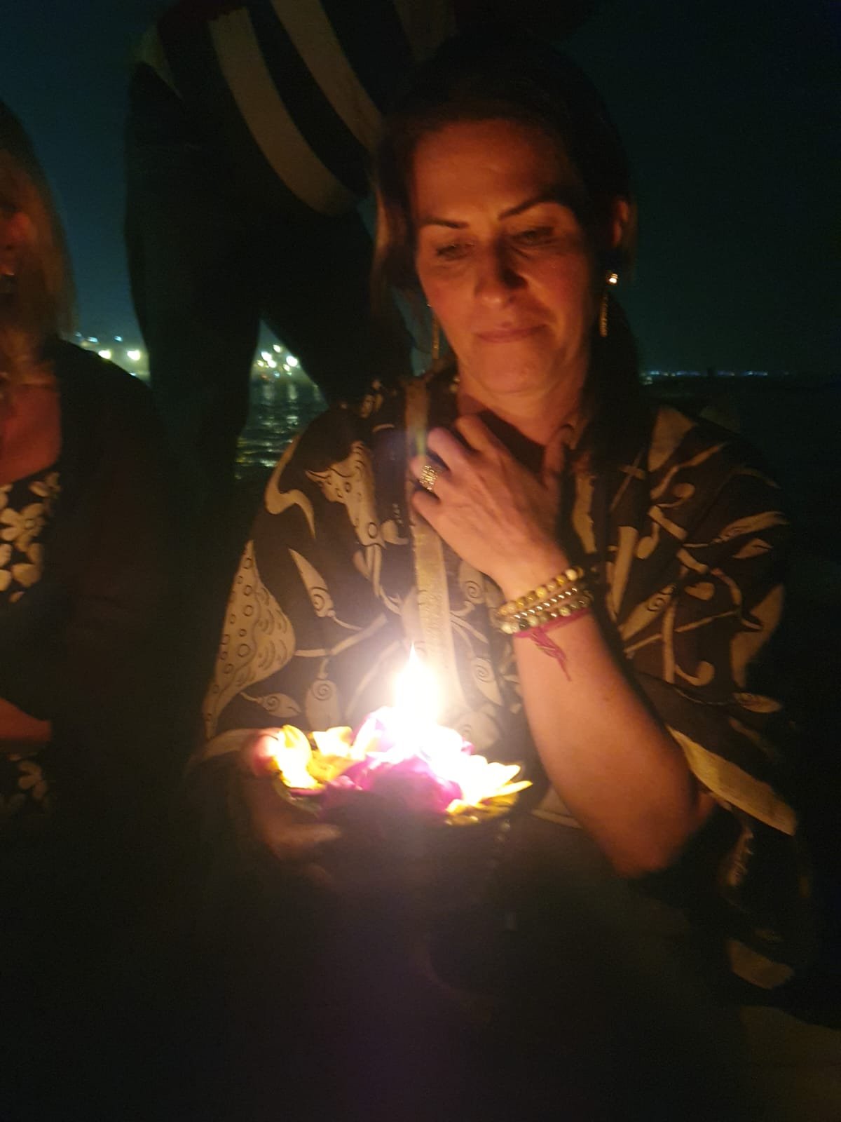 A woman holding a small lit candle in a decorative holder, wearing a patterned shirt with darkness in the background, possibly at night.