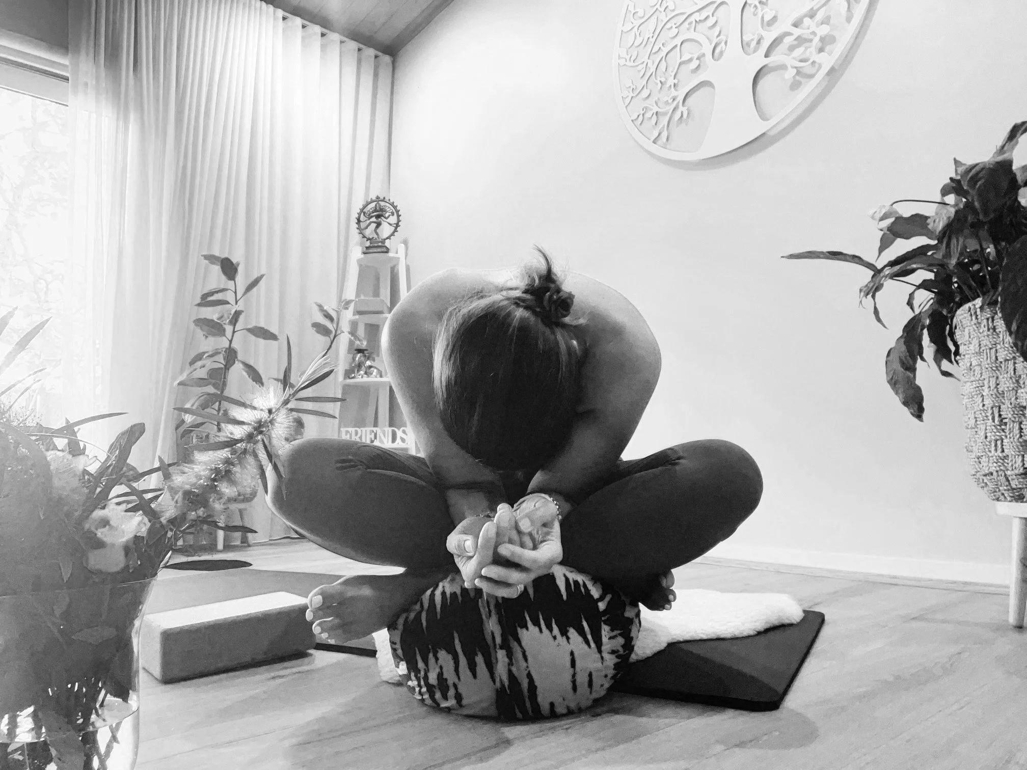 A person practicing yoga indoors, sitting cross-legged with arms wrapped around their feet, head bowed, on a yoga mat near a window with curtains, surrounded by plants and decorative items.