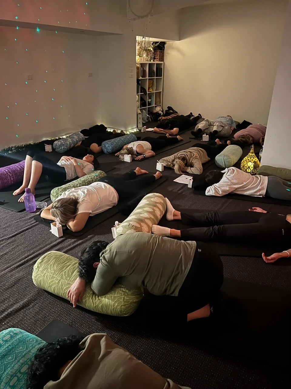 People practicing yoga or meditation on mats with bolsters in a dimly lit room with relaxing decor.