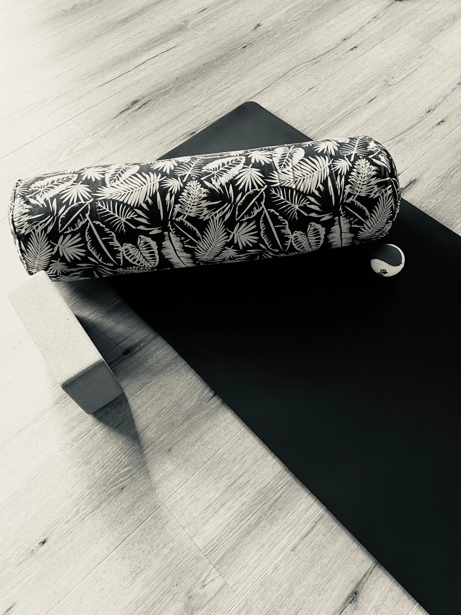 Yoga mat with a black and white tropical leaf pattern, a meditation cushion, and a yin-yang painted ball on a wood floor.