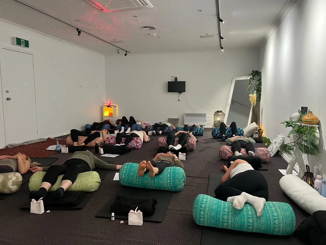 Group of people lying on yoga mats and cushions in a yoga or meditation class, practicing relaxation or yoga poses, inside a spacious room with white walls, decorative plants, and a mirror.