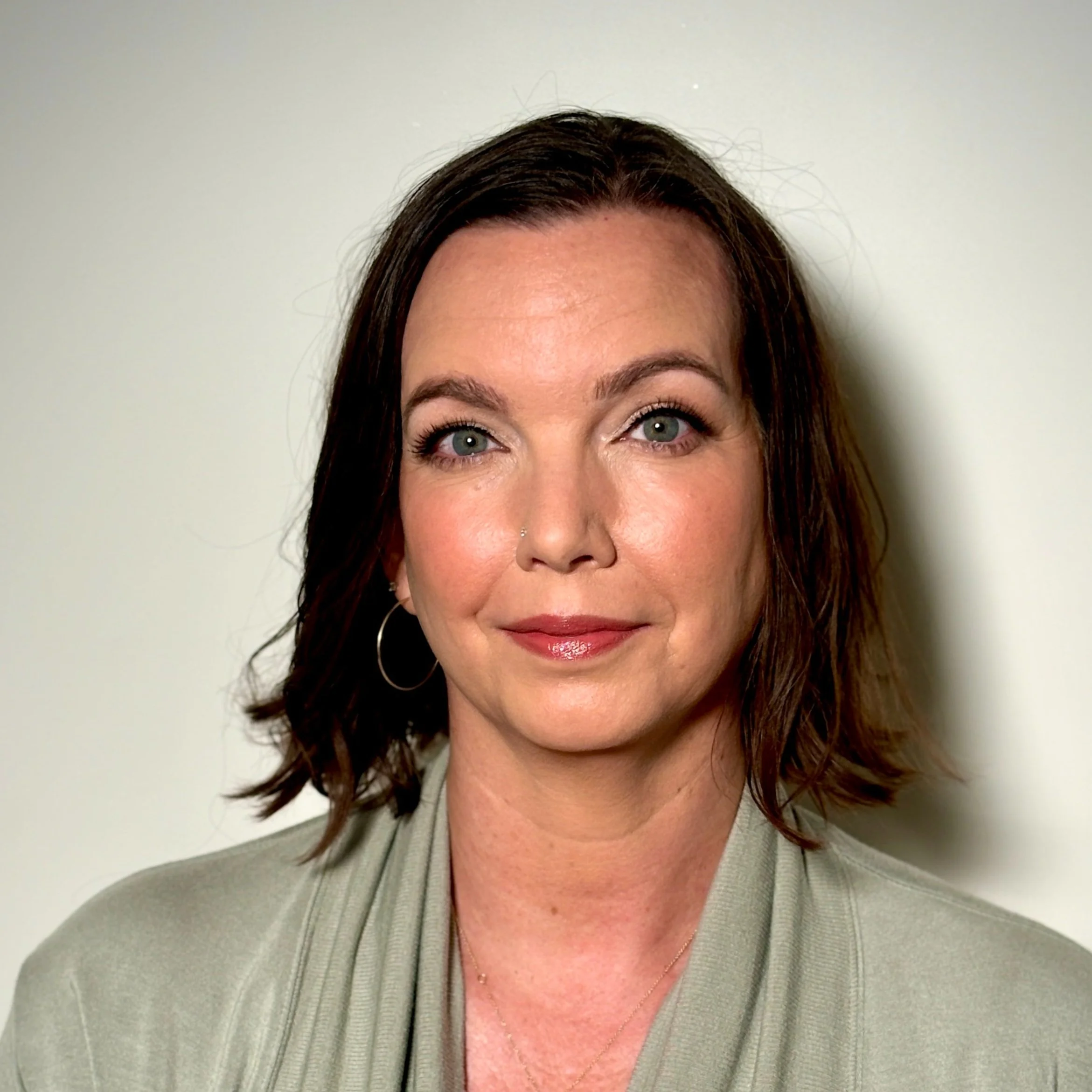 Headshot of a woman with shoulder-length brown hair, green eyes, wearing hoop earrings and a light-colored blazer, smiling softly against a plain white background.