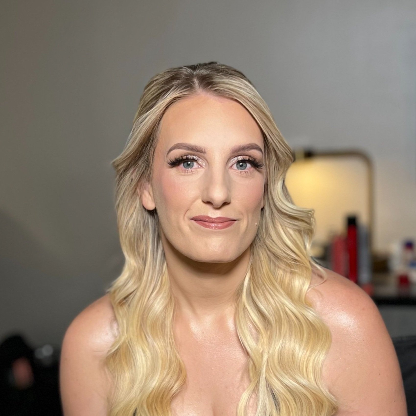 A woman with long, wavy blonde hair and makeup, smiling softly, in an indoor setting with a blurred background.