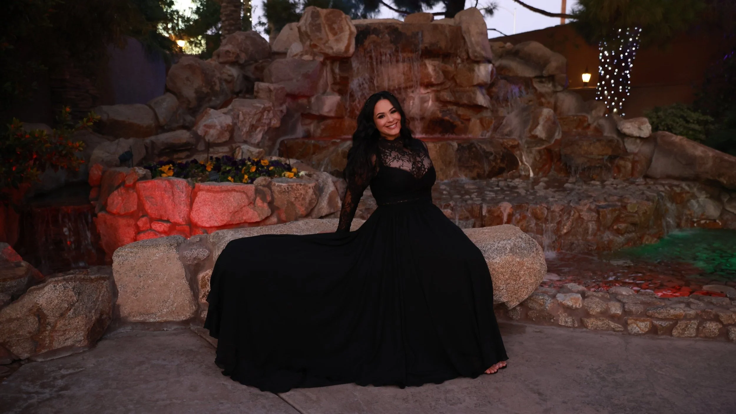 Full glam bridal makeup featured on a bride in a long black lace dress, photographed outdoors at dusk with a rock waterfall backdrop
