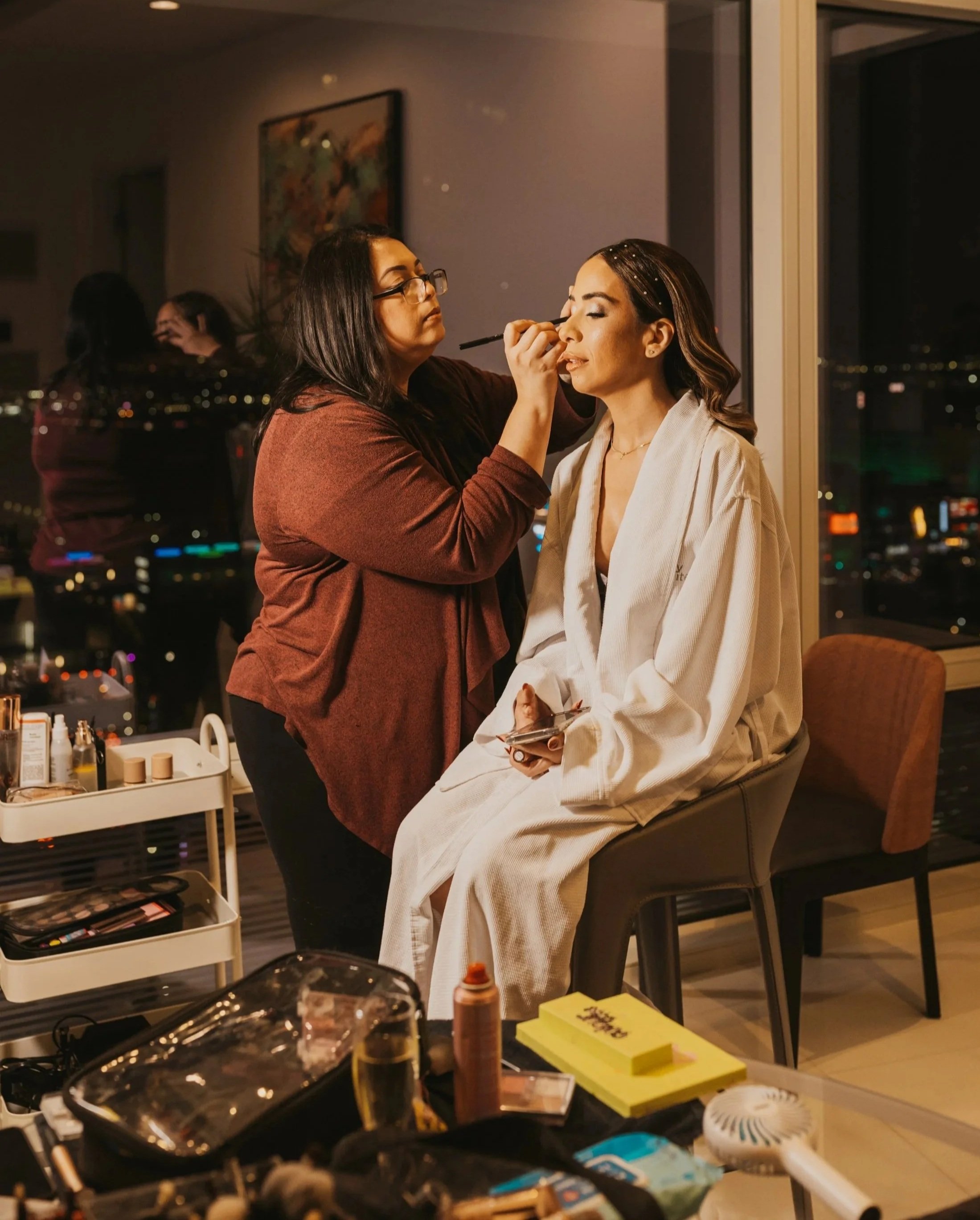 A makeup artist applying makeup to a woman seated on a chair in a room with large windows at night, city lights visible outside.