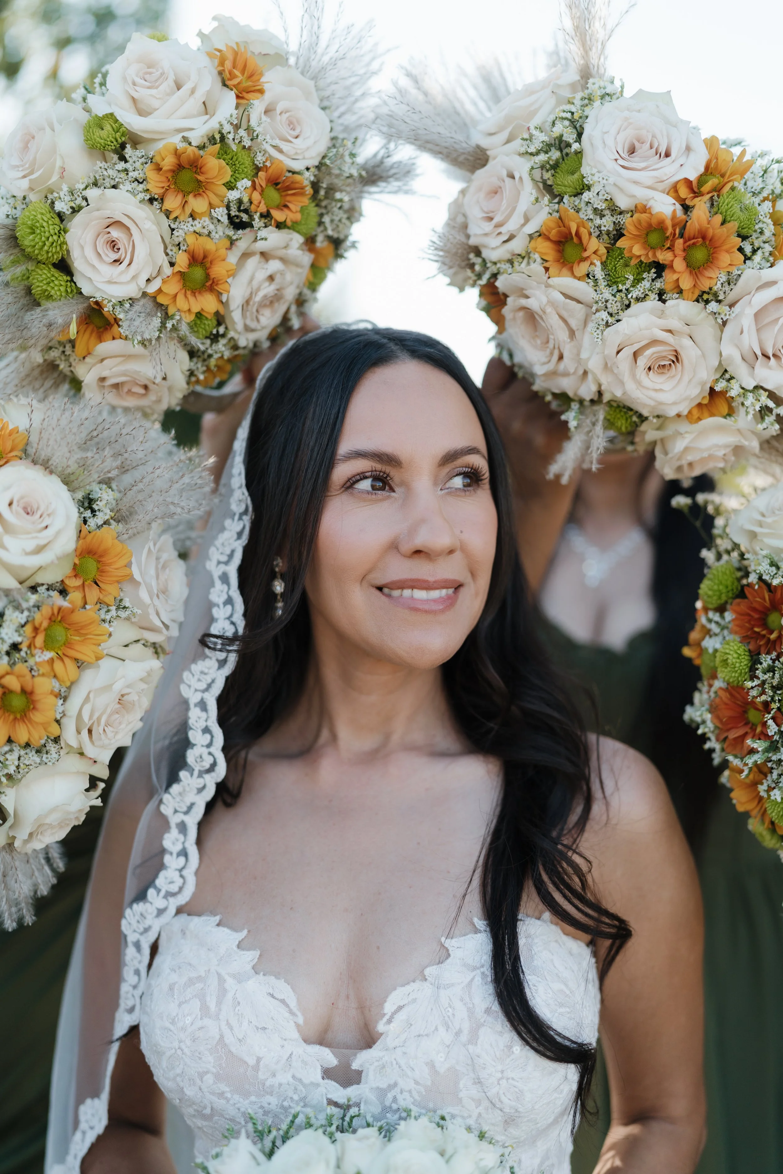 A bride in a white lace wedding dress with a veil, surrounded by large floral arrangements with white roses and orange daisies.