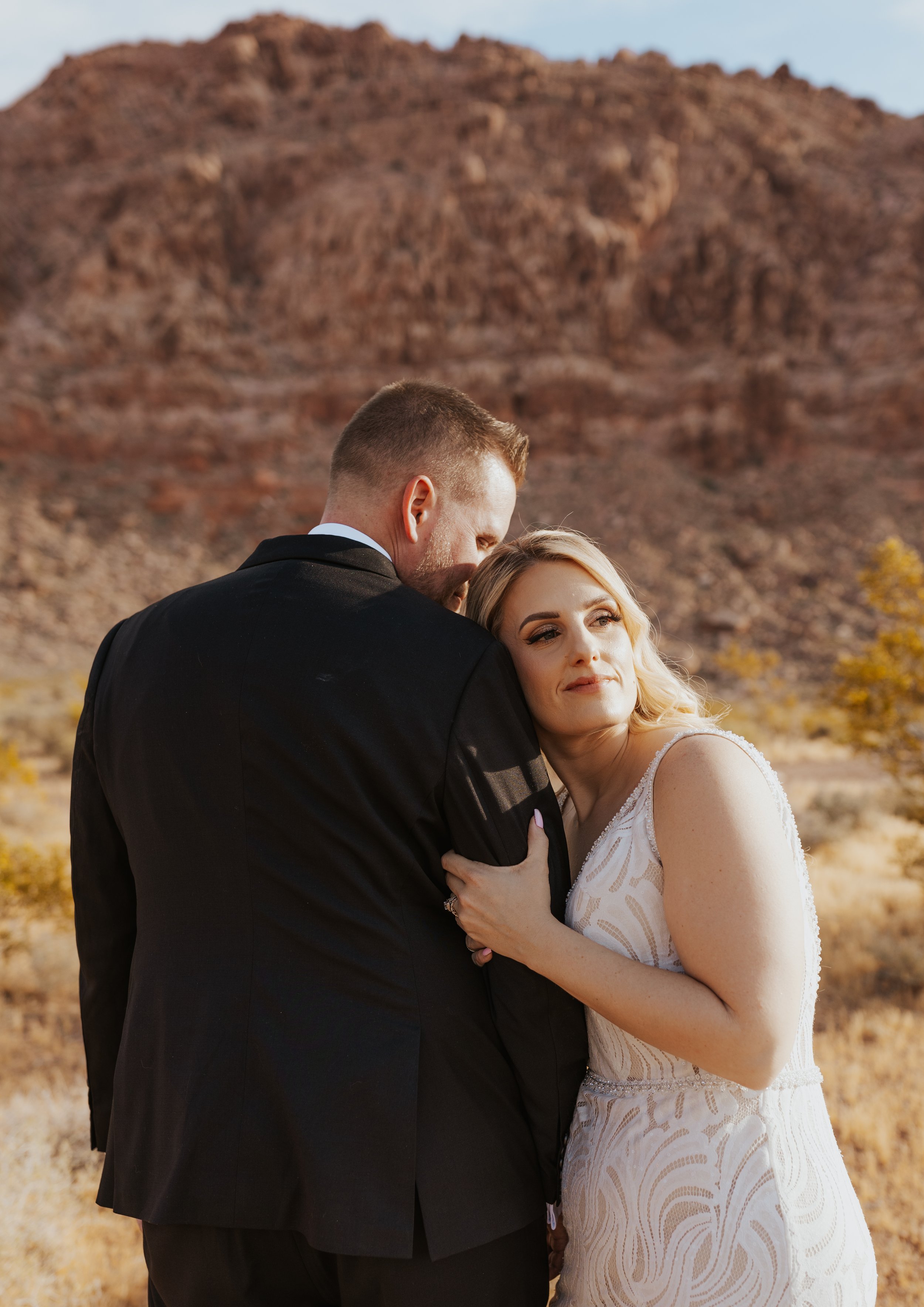 A bride and groom embrace outdoors in front of a rocky mountain during sunset, with the bride resting her head on the groom's shoulder.