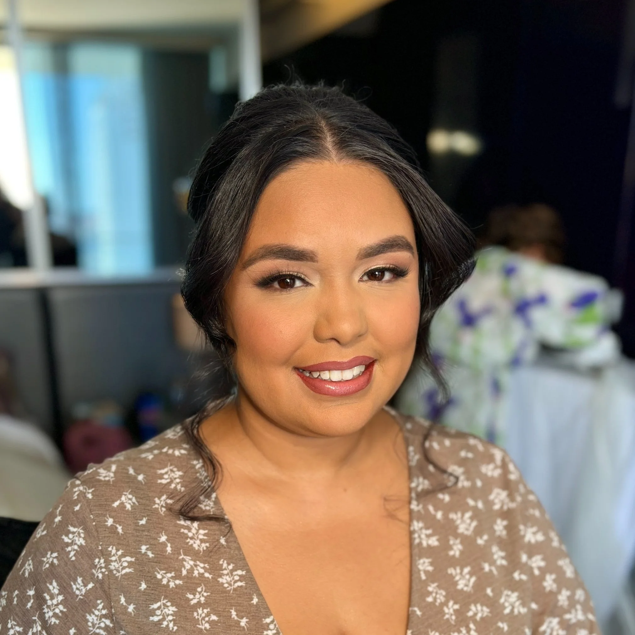 A woman with dark, styled hair and makeup, wearing a brown floral dress, smiling at the camera in an indoor setting.