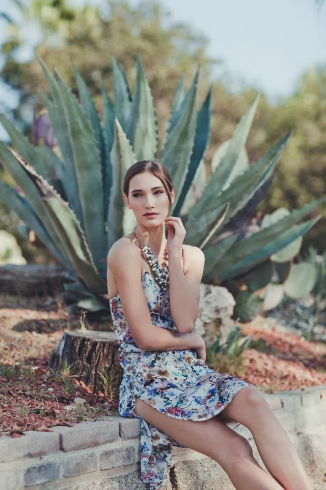 A young woman with brown hair sits on a brick ledge in front of a large agave plant, wearing a floral dress and layered necklace, outdoors on a sunny day.