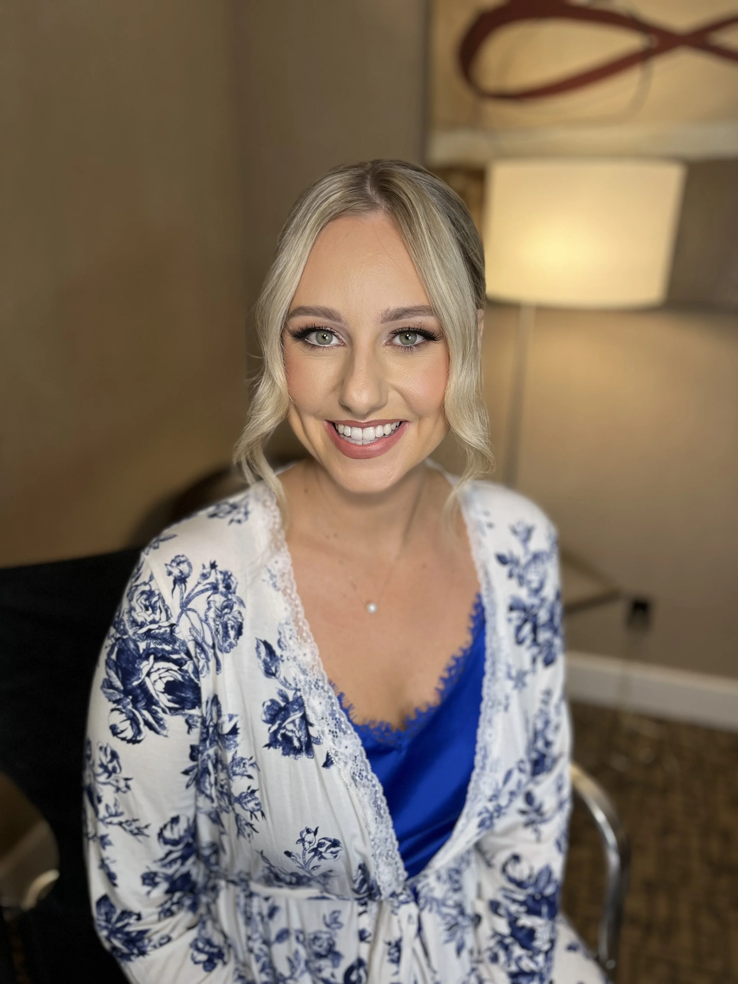 A woman with blonde hair styled in soft waves, wearing a white and blue floral patterned top with lace trim, smiling at the camera inside a room with warm lighting and modern decor.
