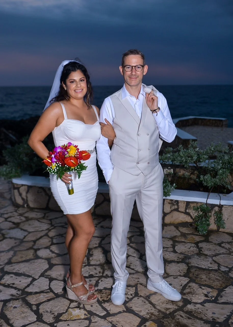 A bride and groom stand together outside near the ocean during sunset. The bride is holding a vibrant bouquet, wearing a white dress and wedge heels, and the groom is dressed in white suit with glasses, carrying a jacket over his shoulder.
