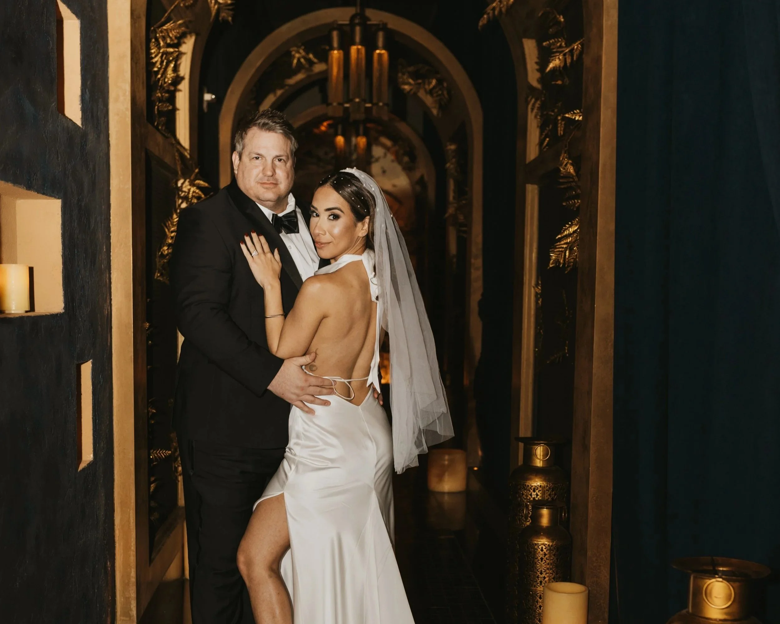 A woman with long, curly black hair and blue eyes wearing a pink dress with silver embellishments, a diamond necklace, and a bracelet, sitting on a chair in a hotel room.