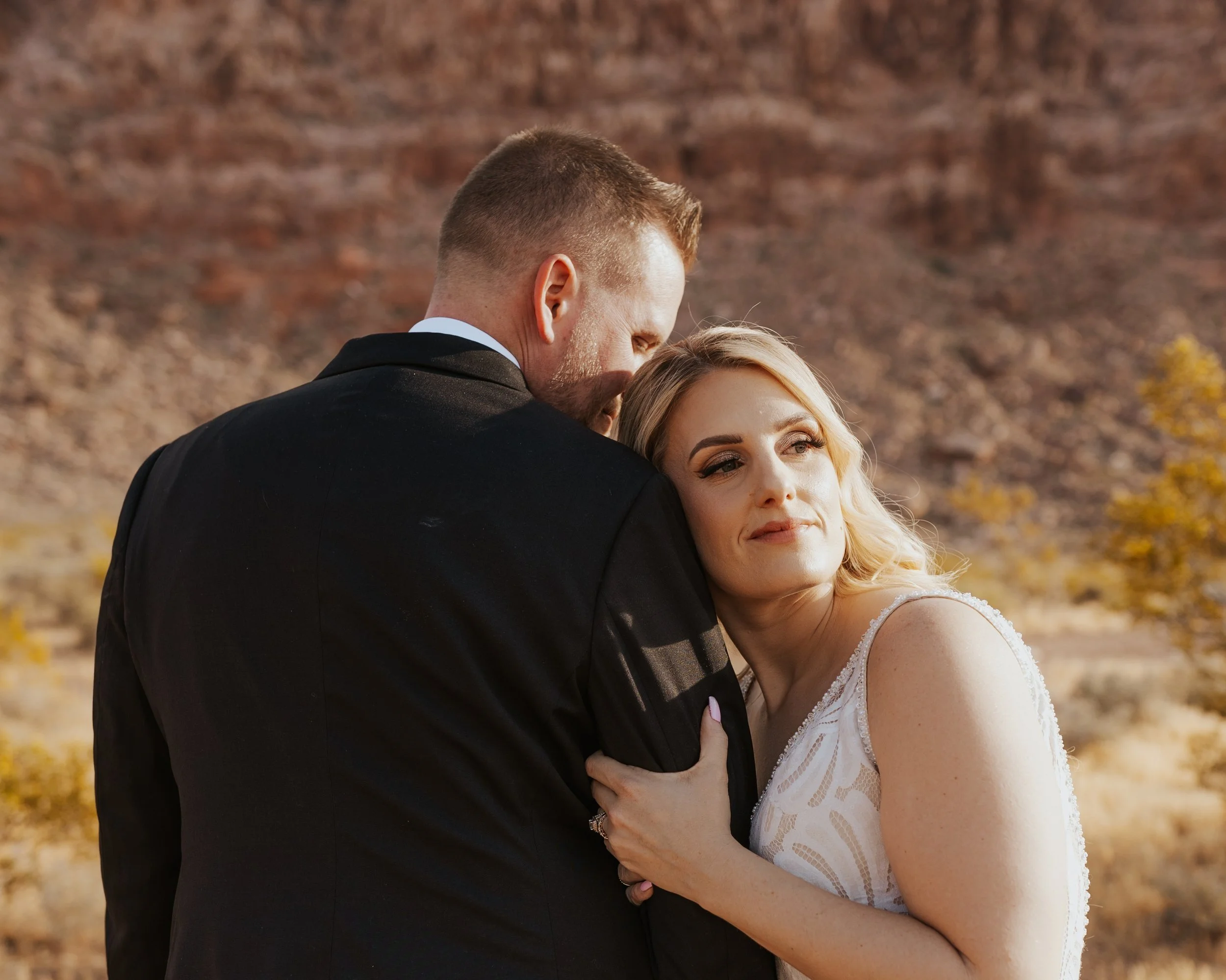 A young woman with dark wavy hair, wearing a black dress with thin straps, smiling in a warmly decorated room with mirrors and floral arrangements.