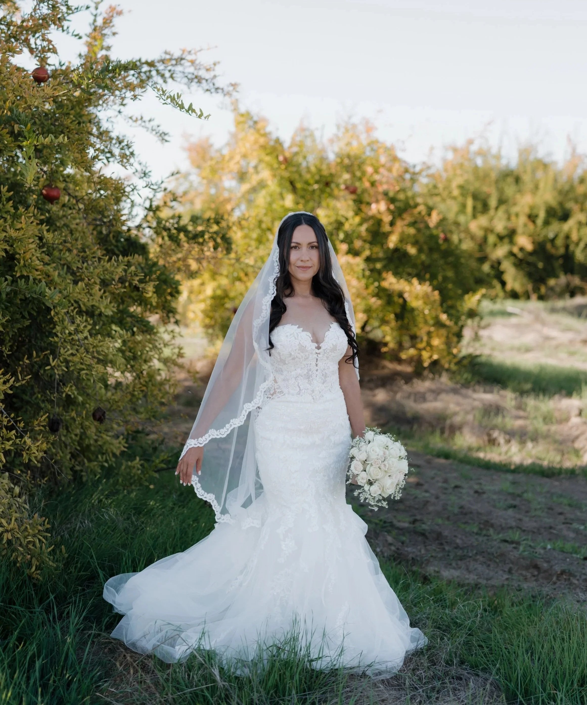 A bride in a white wedding dress holding a bouquet of white roses, standing in a grassy field with trees in the background.
