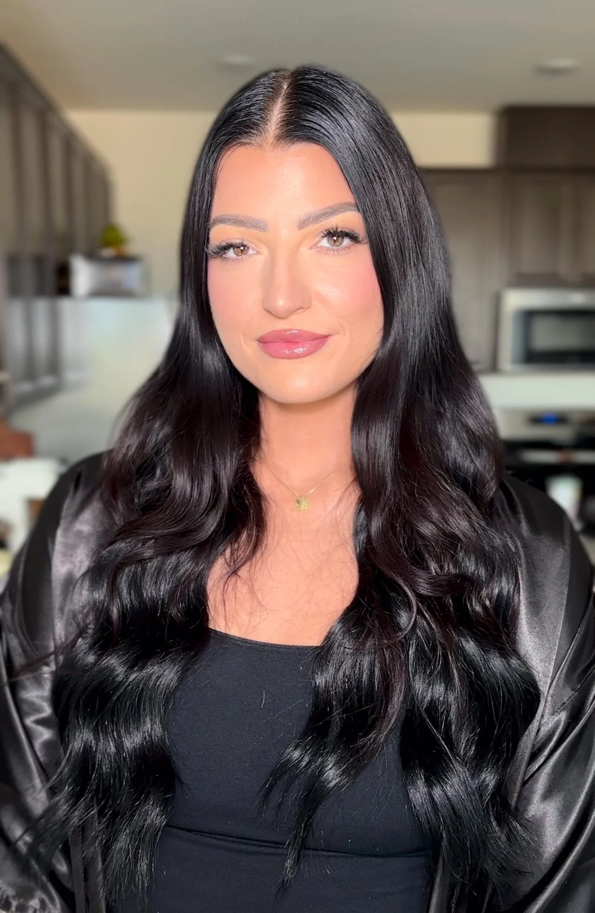 A woman with long, wavy black hair and a natural makeup look, smiling, in a modern kitchen.