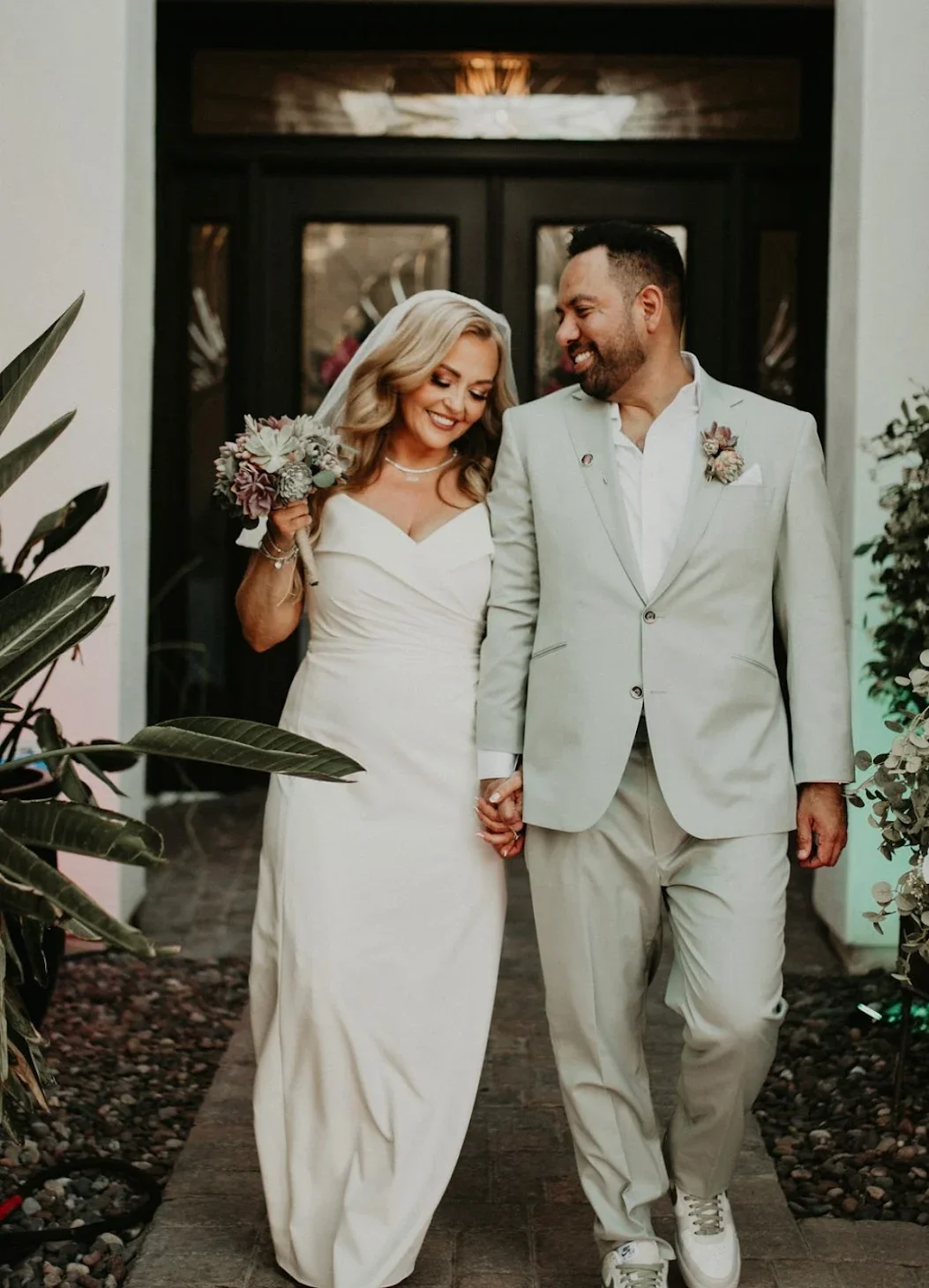 A woman with wavy blonde hair, wearing a black dress with a bow at the front, sitting on a chair in a hotel room. She has makeup and a nose ring, with tattoos on her left arm. Behind her, there is a wall-mounted light, a large landscape painting, a black rolling suitcase, and a patterned carpet.