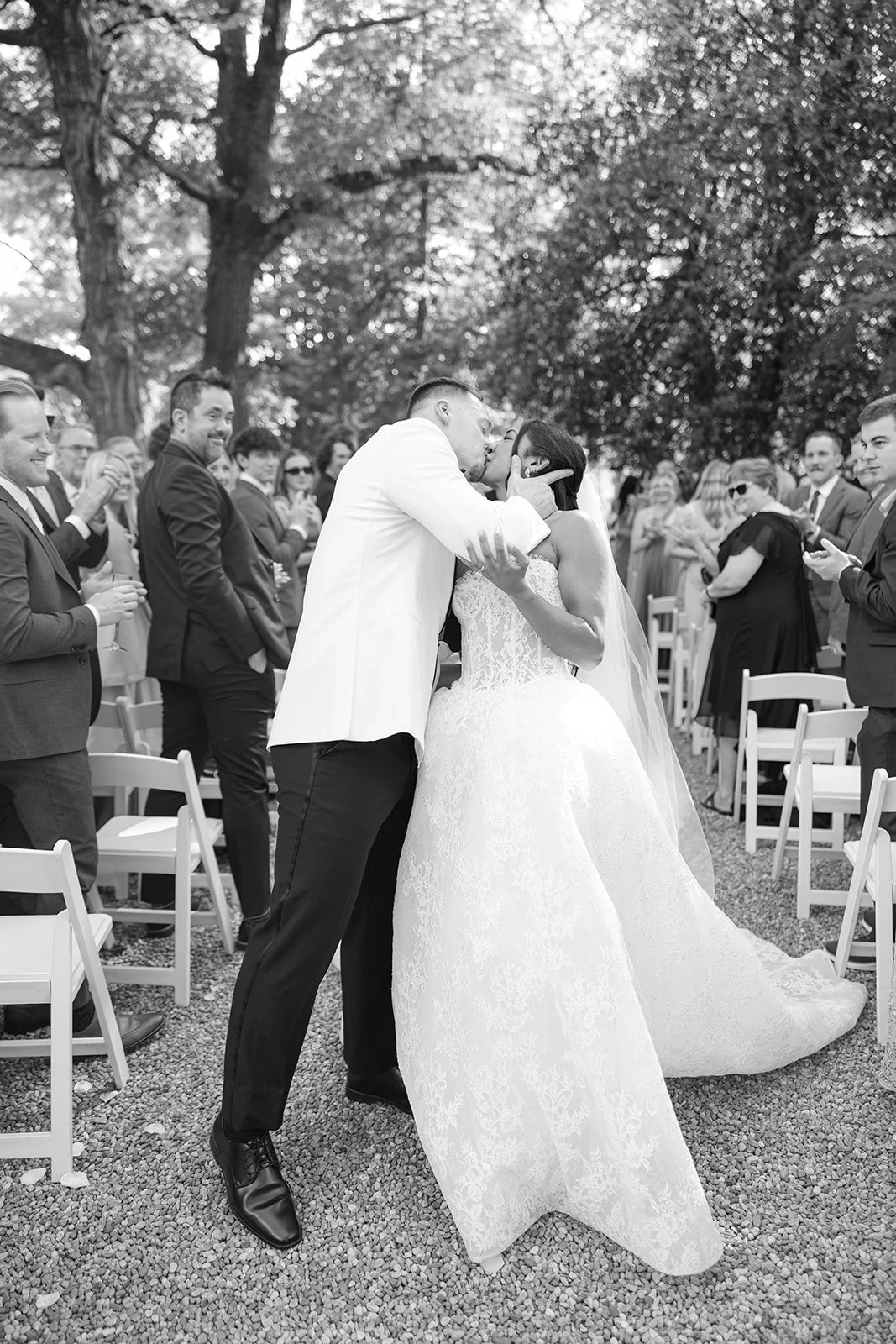 Black and white photo of a bride and groom sharing a kiss at their outdoor wedding ceremony, with guests watching and clapping in the background.