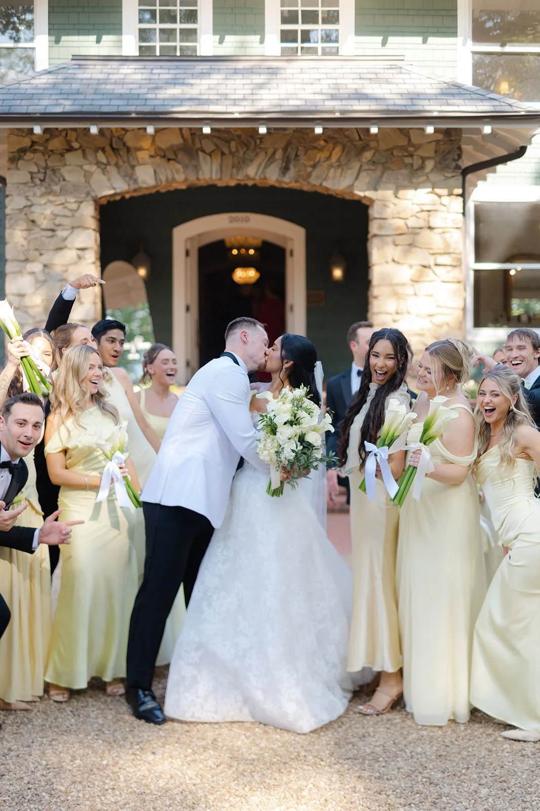 A wedding celebration with a bride and groom kissing in front of a wedding party. The bride holds a bouquet of white flowers, and several bridesmaids hold calla lilies. The wedding party is standing outside a house with stone and green siding.