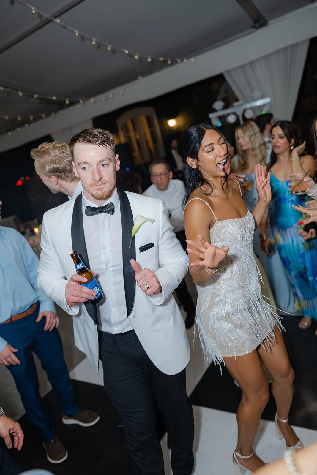 People dancing at a wedding reception, with a man in a white tuxedo holding a beer and a woman in a sparkly dress dancing and smiling.