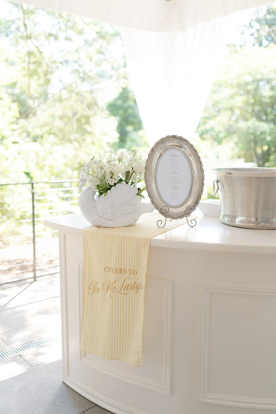 Decorative table with a white swan-shaped vase holding white flowers, a silver picture frame, and a silver ice bucket, set against a bright window with greenery outside. A yellow striped cloth with the words 'Cheers to The McLarty's' hangs from the t