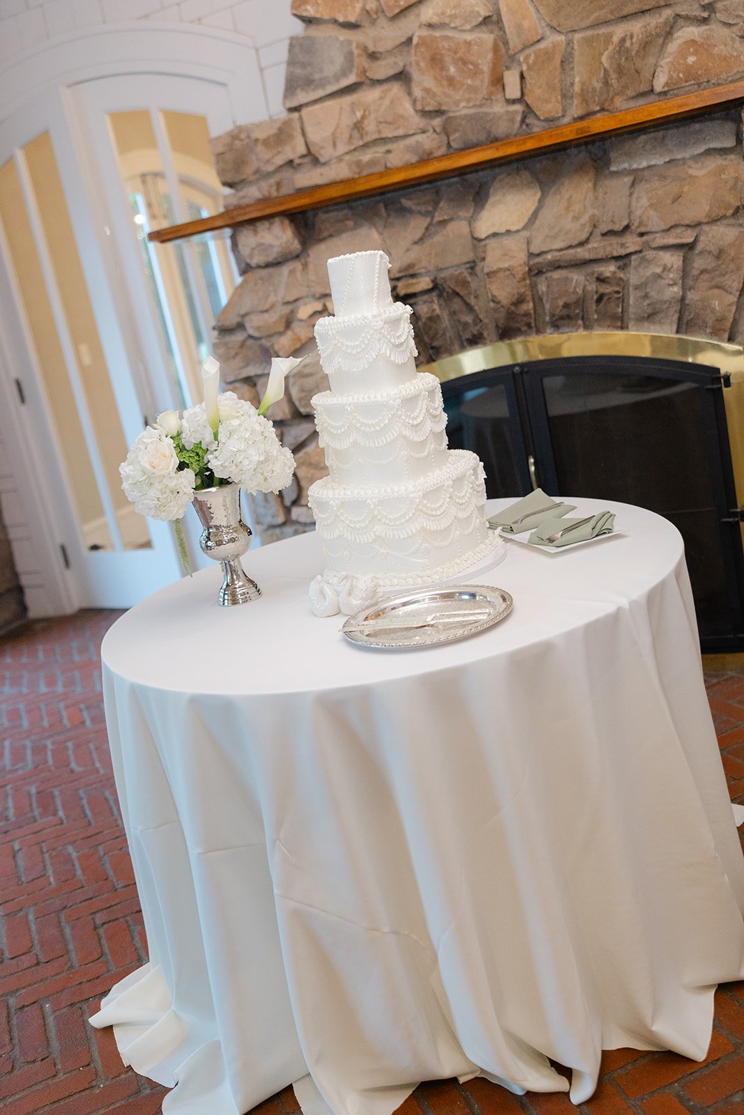 Elegant white wedding cake with multiple tiers decorated with intricate icing patterns, placed on a table with a white cloth. The table is next to a vase of white flowers and silver serving tray, set against a rustic stone fireplace