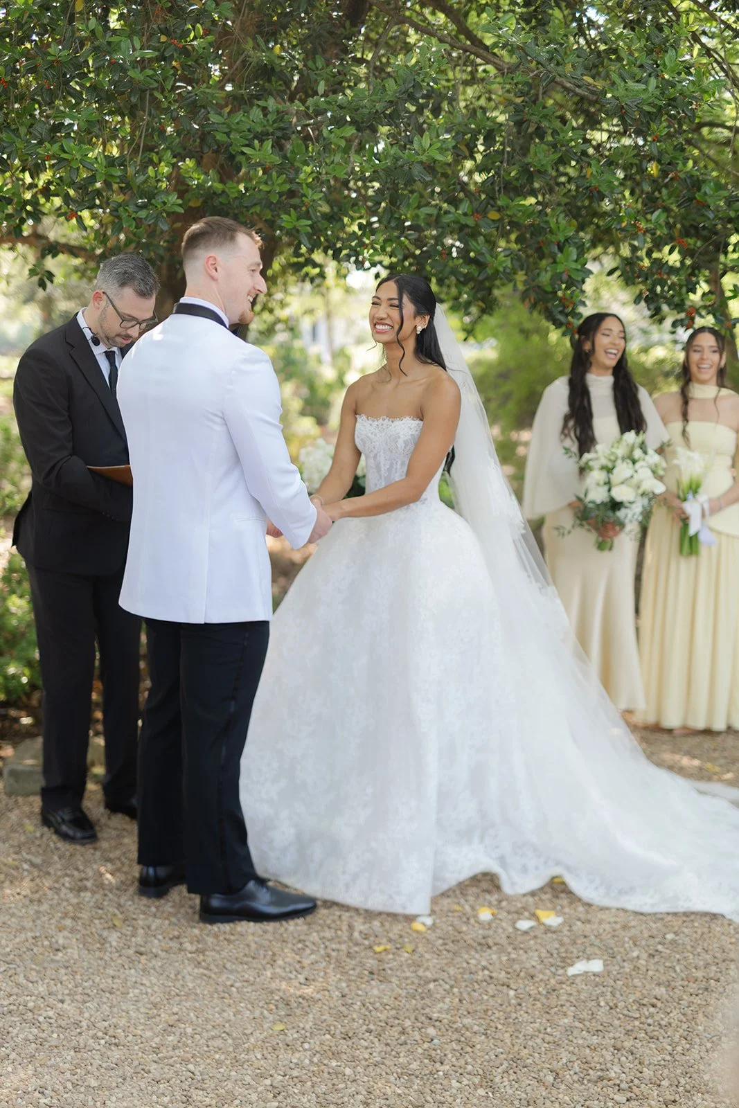 A bride and groom exchanging vows outdoors under a tree, with bridesmaids holding bouquets in the background.