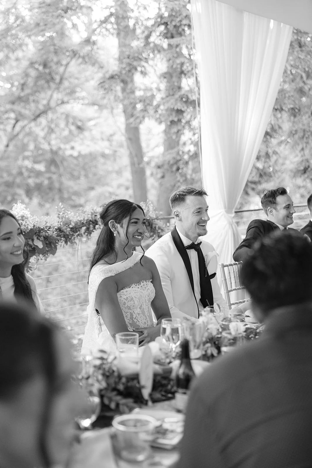Black and white photo of a wedding reception with the bride and groom seated at the table, smiling. The bride wears a lace wedding dress, and the groom is in a tuxedo. Other guests are also visible, enjoying the celebration outdoors with trees in the