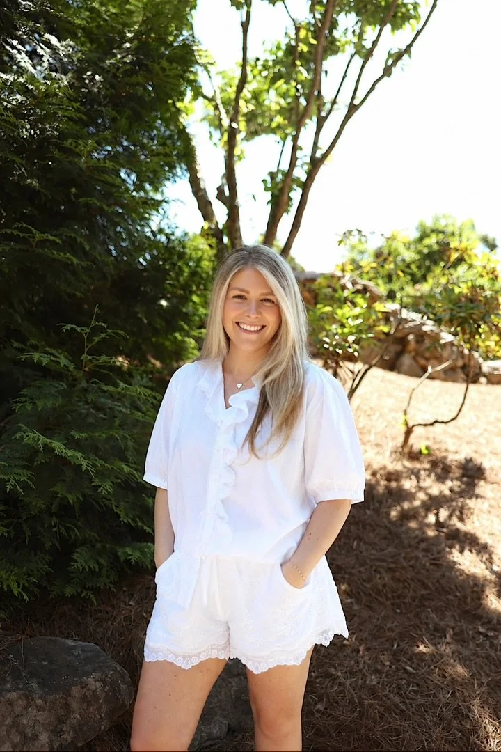 A young woman with blonde hair, wearing a white blouse and white shorts, smiling and standing outdoors among trees and greenery.